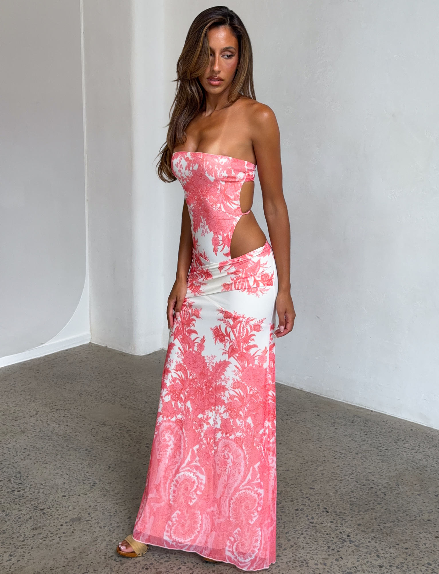 A woman wears the TIGERMIST POPPI MAXI DRESS - CORAL TAPESTRY, a strapless long white dress with bold pink floral and coral tapestry print plus side cutouts, standing indoors on a polished concrete floor against a white wall.