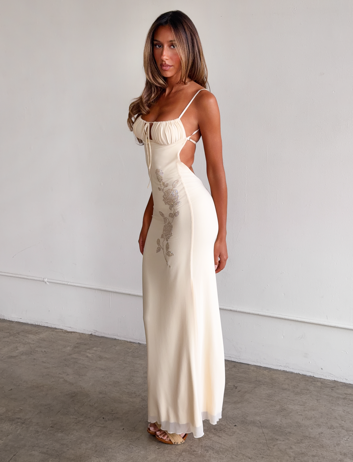 A woman with long, wavy hair poses indoors against a plain wall, wearing the TIGERMIST OPHELIA MAXI DRESS in cream, featuring adjustable straps and delicate floral embroidery. She gazes slightly to the side.