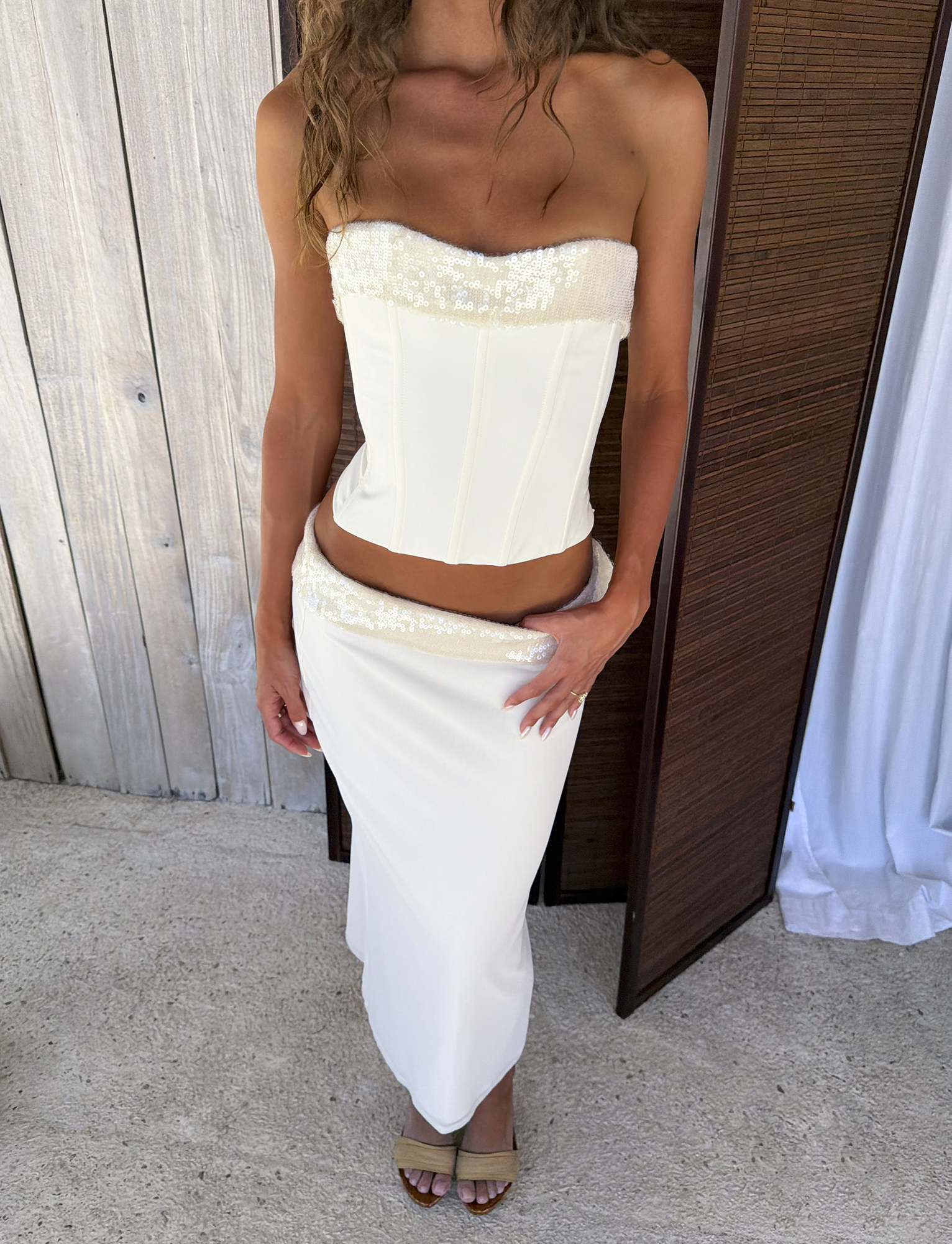 A woman stands indoors in the TIGERMIST CHARLYZE BUSTIER in cream, a strapless top with sequin paneling, paired with a matching long skirt and open-toe heels. Wooden panels and white curtains are in the background.