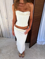 A woman stands indoors in the TIGERMIST CHARLYZE BUSTIER in cream, a strapless top with sequin paneling, paired with a matching long skirt and open-toe heels. Wooden panels and white curtains are in the background.