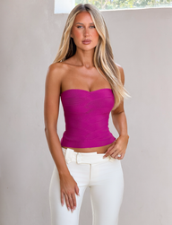 A woman with long blonde hair models the CL - NIYA METALLIC BANDAGE TOP by Tiger Mist, a strapless, fitted magenta piece, paired with high-waisted white pants. She stands indoors by a light wall under natural window light.