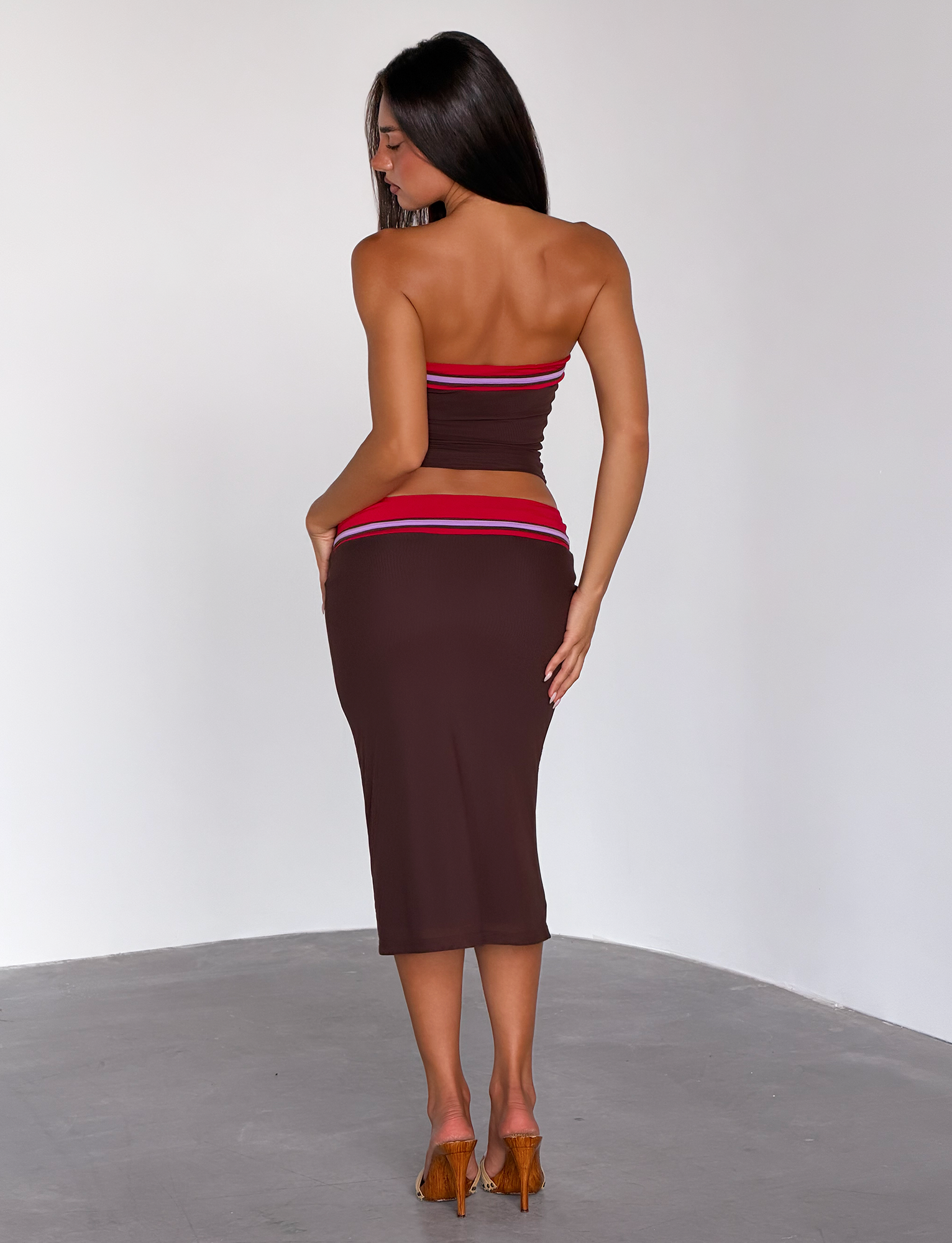 A woman with long dark hair stands barefoot on a gray floor, facing away. She wears the TIGERMIST SERAH DIAMANTE MIDI SKIRT in chocolate and a matching 90’s-inspired crop top. The background is plain white.