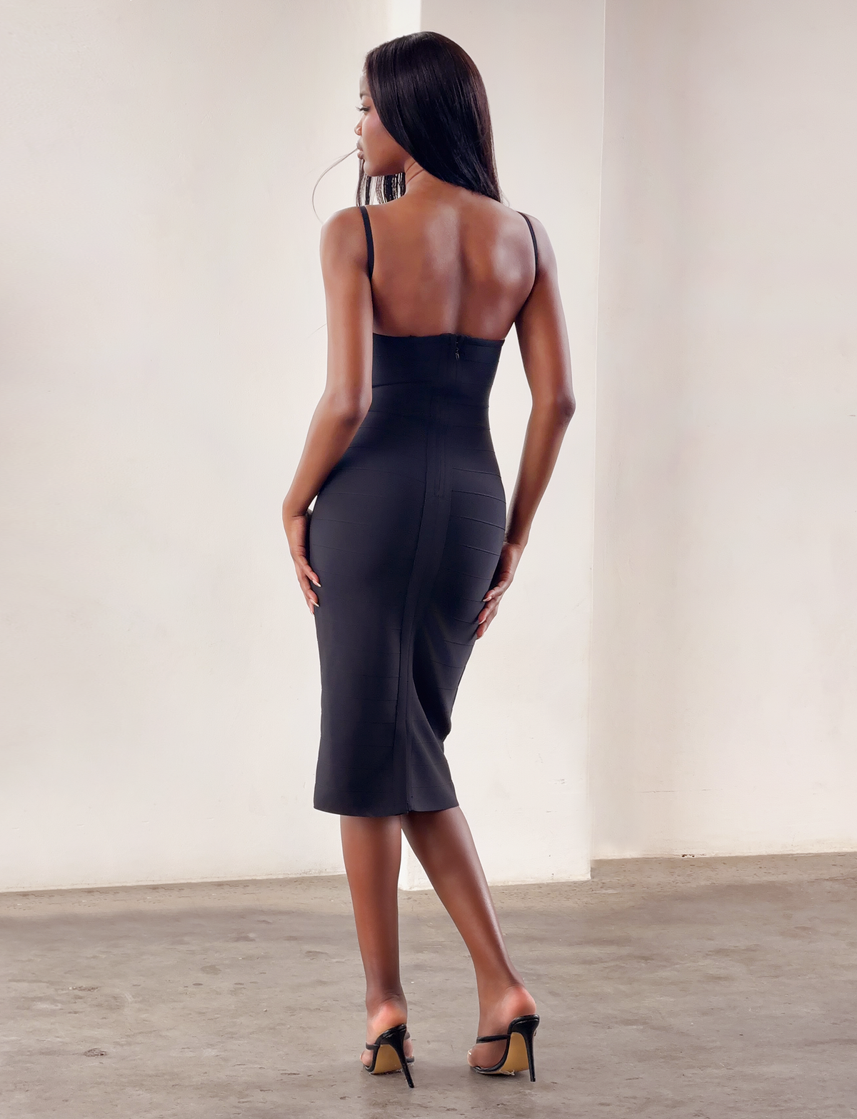 A woman stands indoors facing away, wearing the MIDORI BANDAGE MIDI DRESS in black by TIGERMIST. The fitted, bodycon dress accentuates her silhouette, and she pairs it with black high-heeled sandals. Her long hair is straight as she poses with one leg slightly bent.
