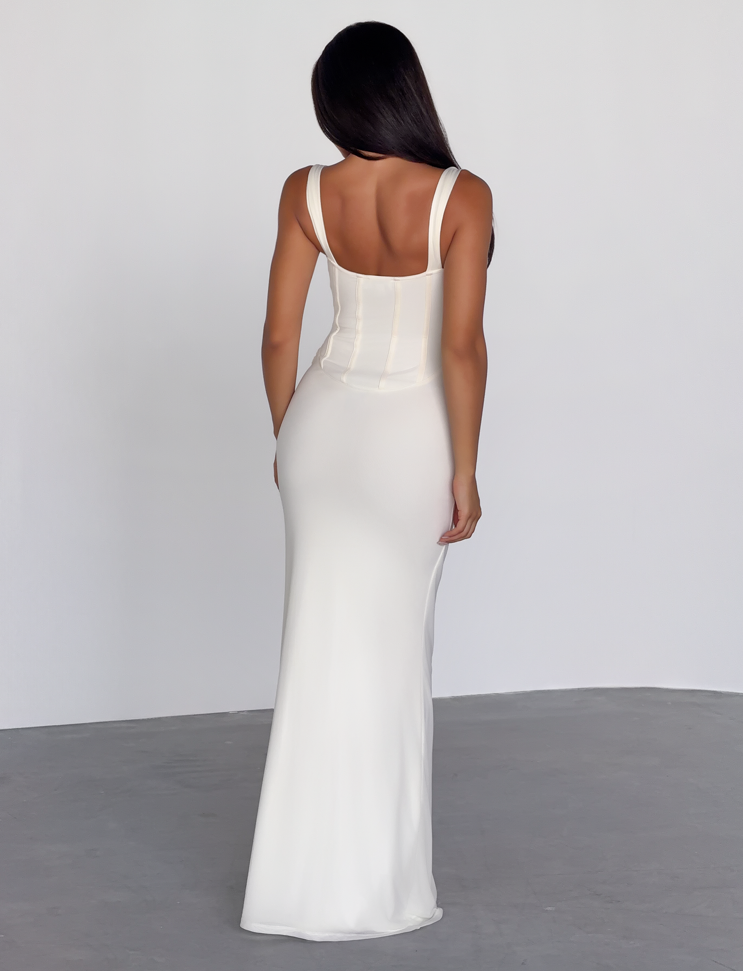 A woman with long, dark hair stands in a minimalistic room, facing away from the camera, wearing the TIGERMIST GINNY MAXI DRESS in off white featuring a corseted bodice, floor-length hem, and elegant draped mesh detail.