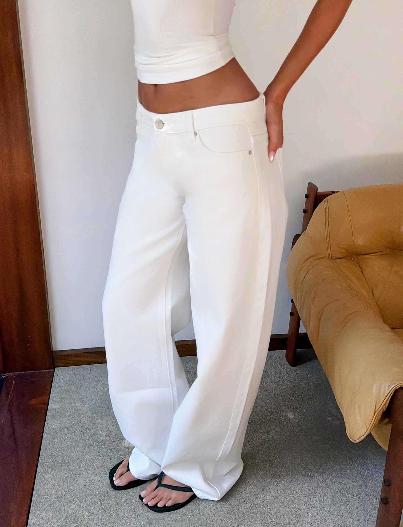 Wearing a fitted white sleeveless top, TIGERMIST MALIYA JEAN - WHITE, and black sandals, a person stands indoors next to a tan armchair with one hand on their hip.
