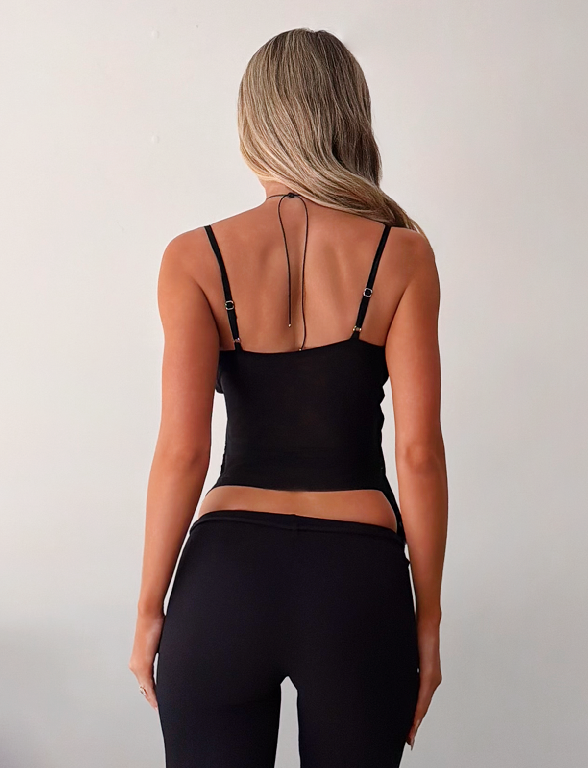A woman with long, wavy blonde hair faces away, wearing the TIGERMIST ALDEANA TOP in black and black pants against a plain light-colored background.