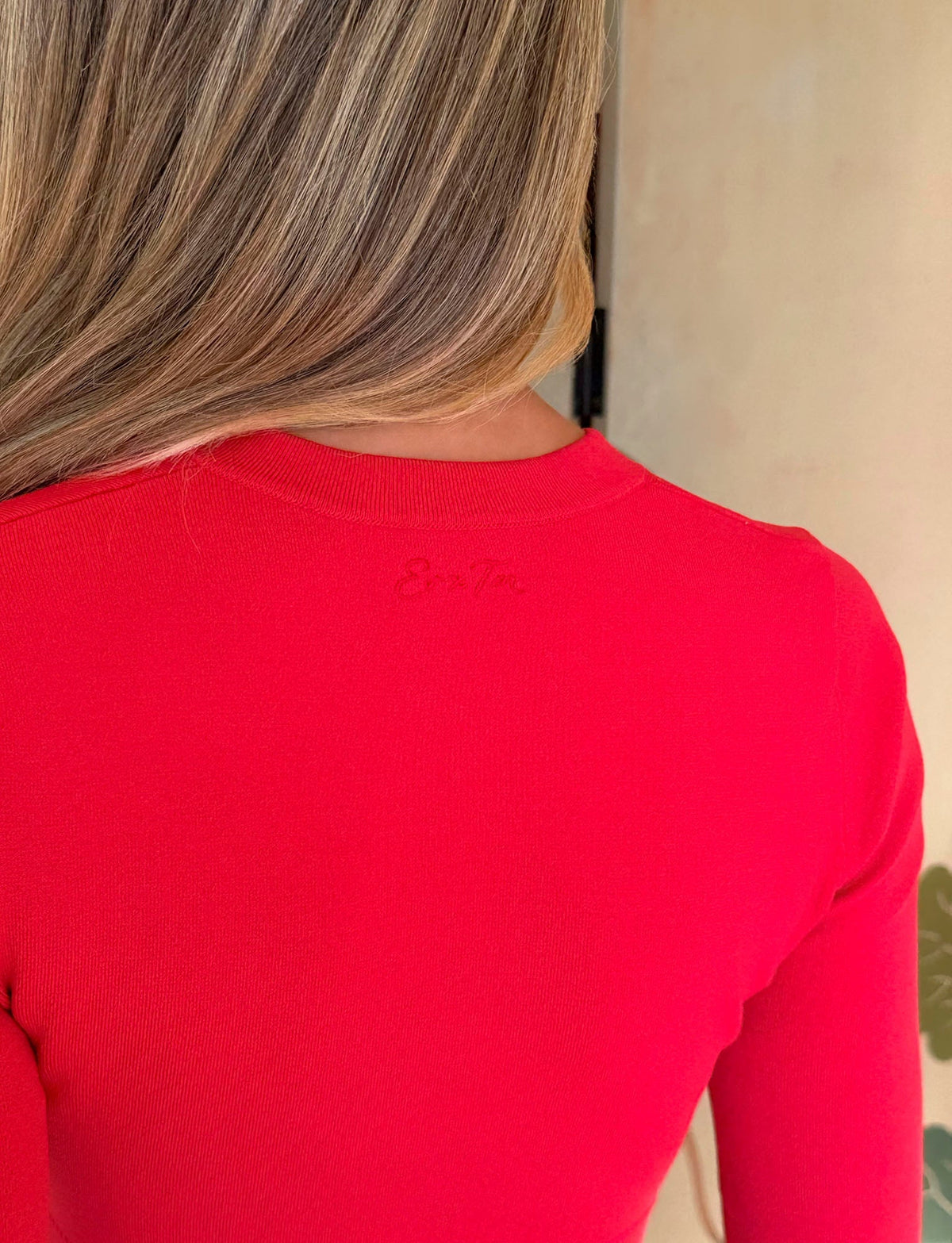 A person with long, highlighted hair is seen from behind, wearing the TIGERMIST DAMARIS CARDIGAN in red, which features subtle text near the collar.