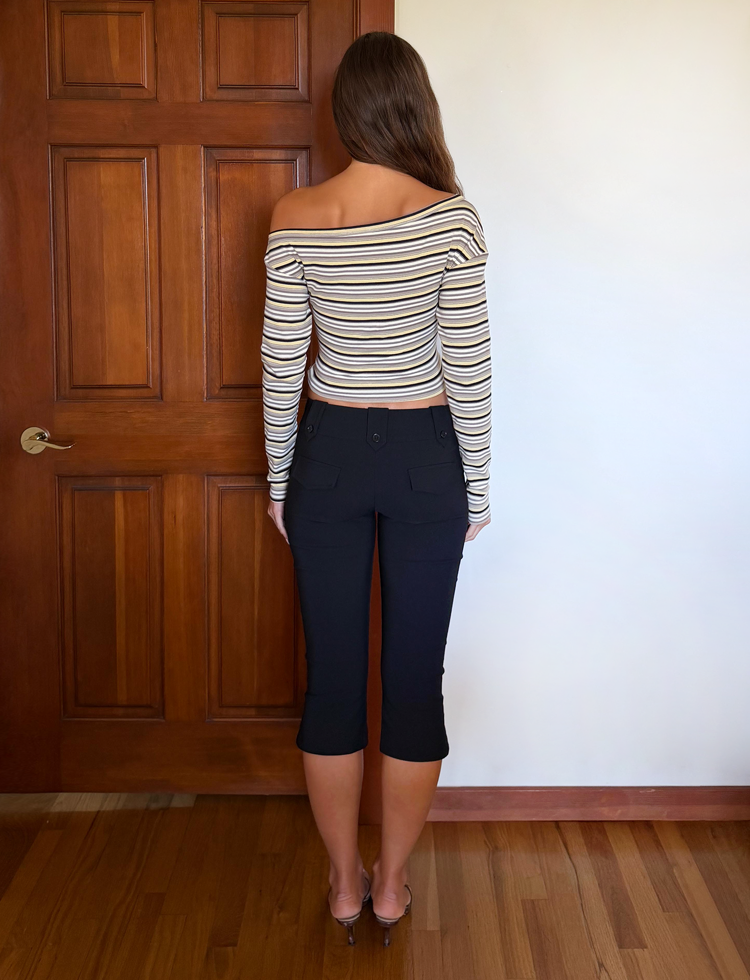 A woman with long brown hair stands indoors facing away, wearing the TIGERMIST KIERSTEN SCARF TOP in multistripe and black cropped pants near a brown door and light wall.