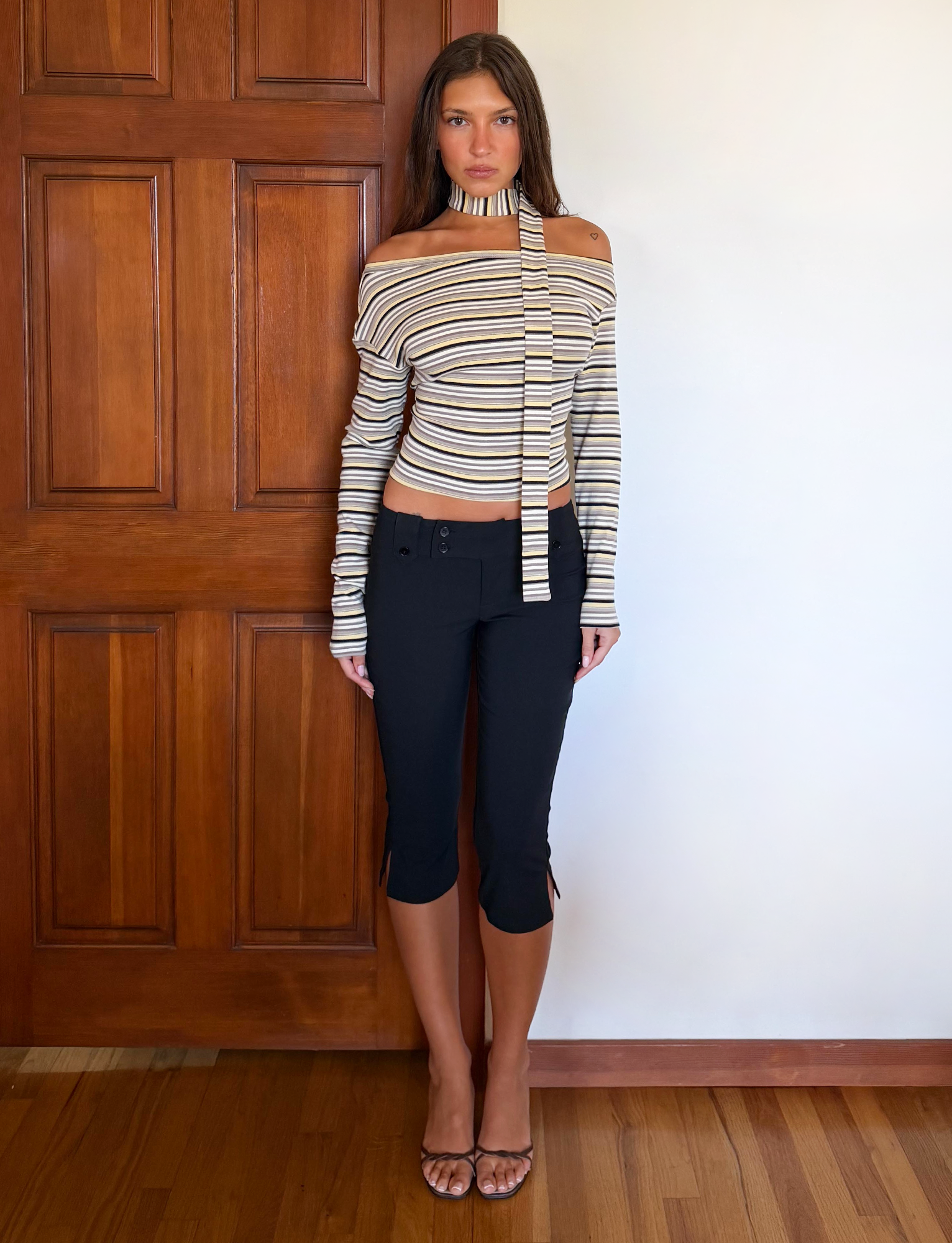 A woman with long brown hair and a neutral expression stands indoors by a wooden door, wearing the TIGERMIST KIERSTEN SCARF TOP - MULTISTRIPE with black cropped pants and clear heeled sandals.