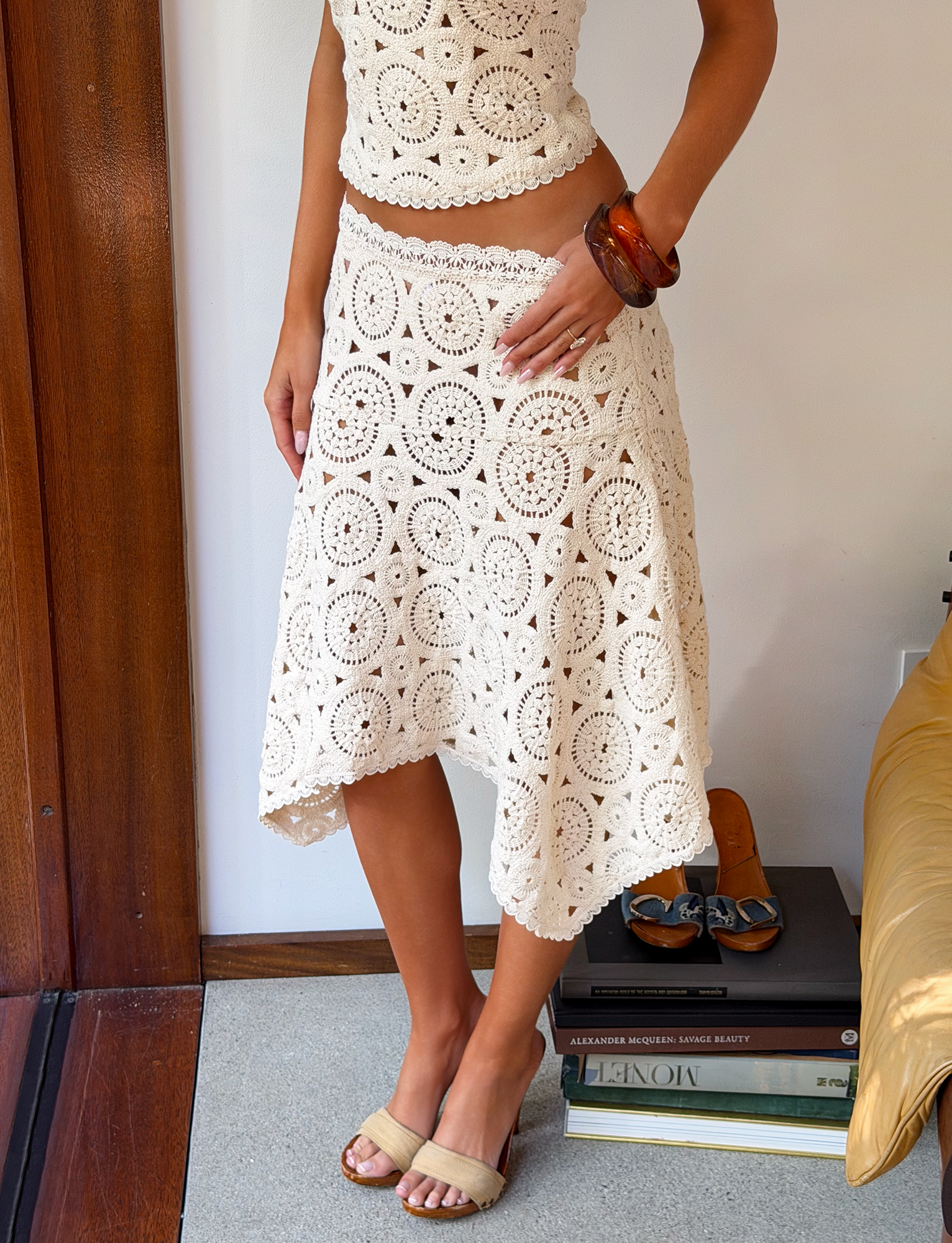 A person stands indoors wearing the TIGERMIST SYNTHIA CROCHET MIDI SKIRT in cream, featuring circular crochet patterns. Barefoot beside books and heels, they accessorize with a brown bangle; their face is not shown.