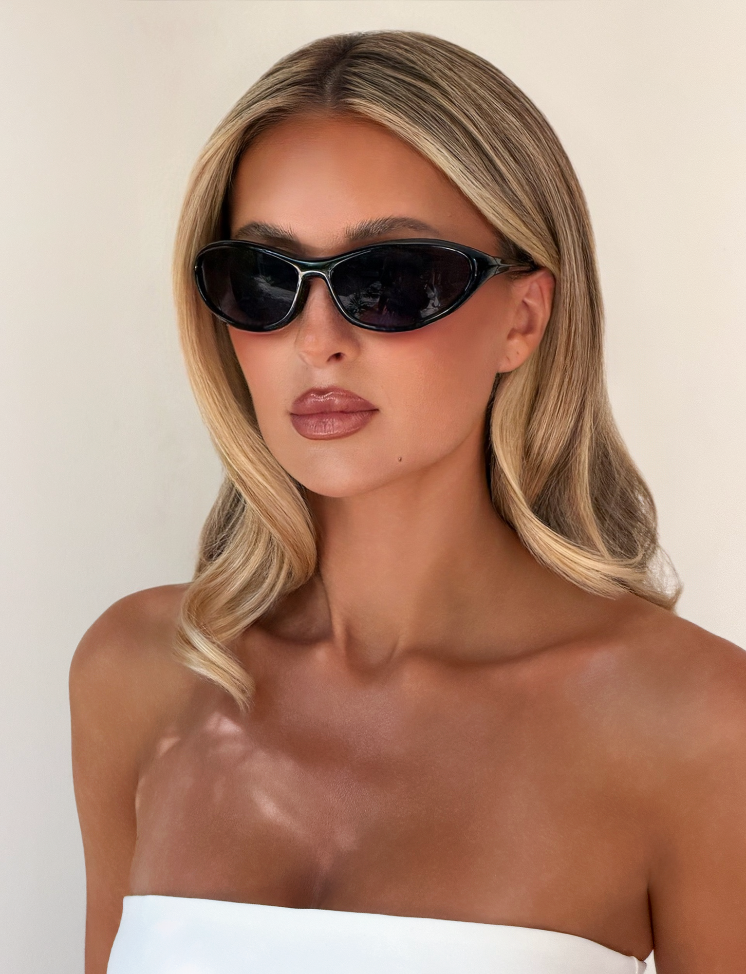 A woman with long, blonde hair models the TIGERMIST RIVAH FASHION GLASSES - BLACK, featuring oval frames and reflective black lenses. She wears a white strapless top and poses against a plain light background with a neutral expression.