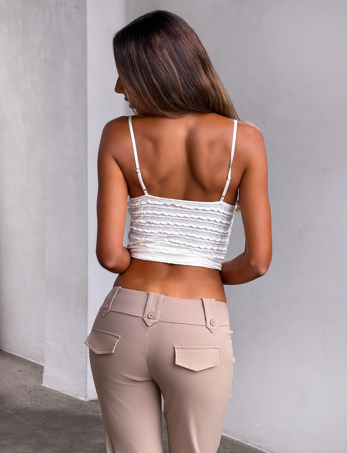 A woman with long brown hair stands with her back to the camera, wearing the TIGERMIST ZORA TOP in cream, featuring adjustable shoulder straps, paired with high-waisted beige pants against a plain light wall and floor.