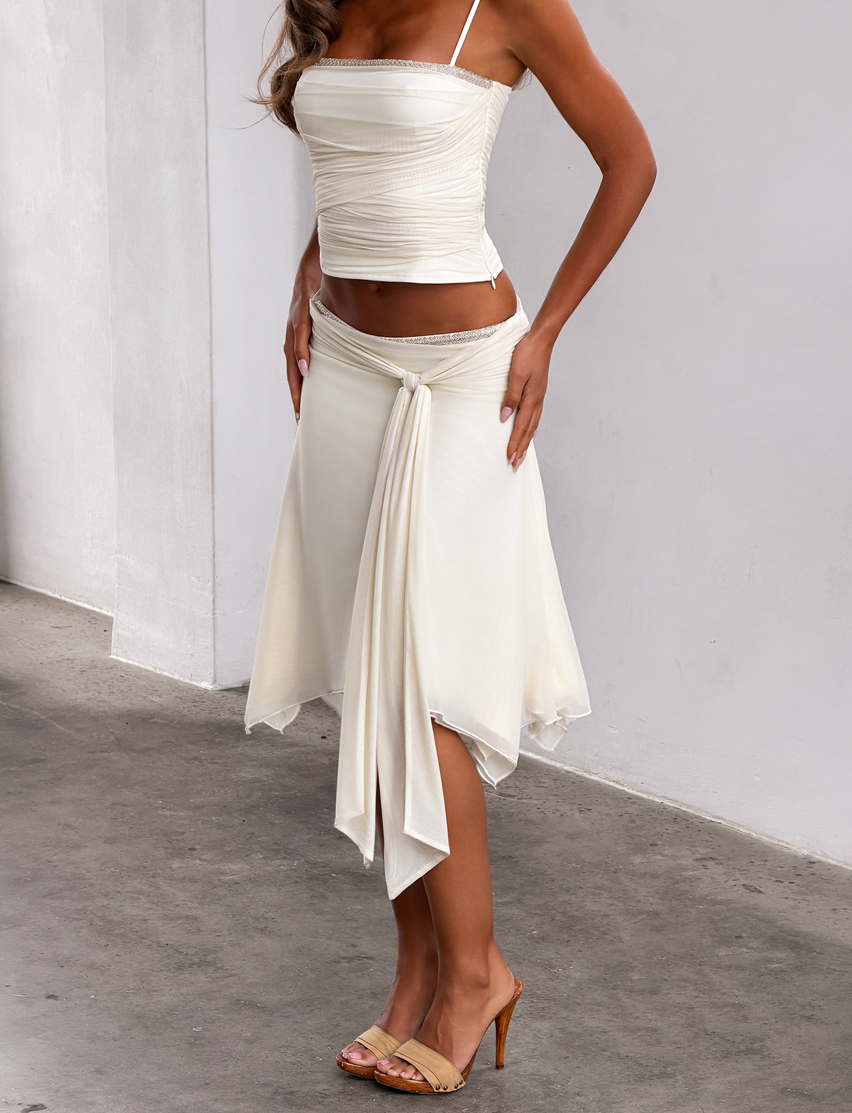 A woman wearing a white sleeveless top and the TIGERMIST JUNA MIDI SKIRT in cream, featuring an elegant mesh fabric with draped detailing, stands on a concrete floor by a white wall, paired with tan high-heeled sandals.