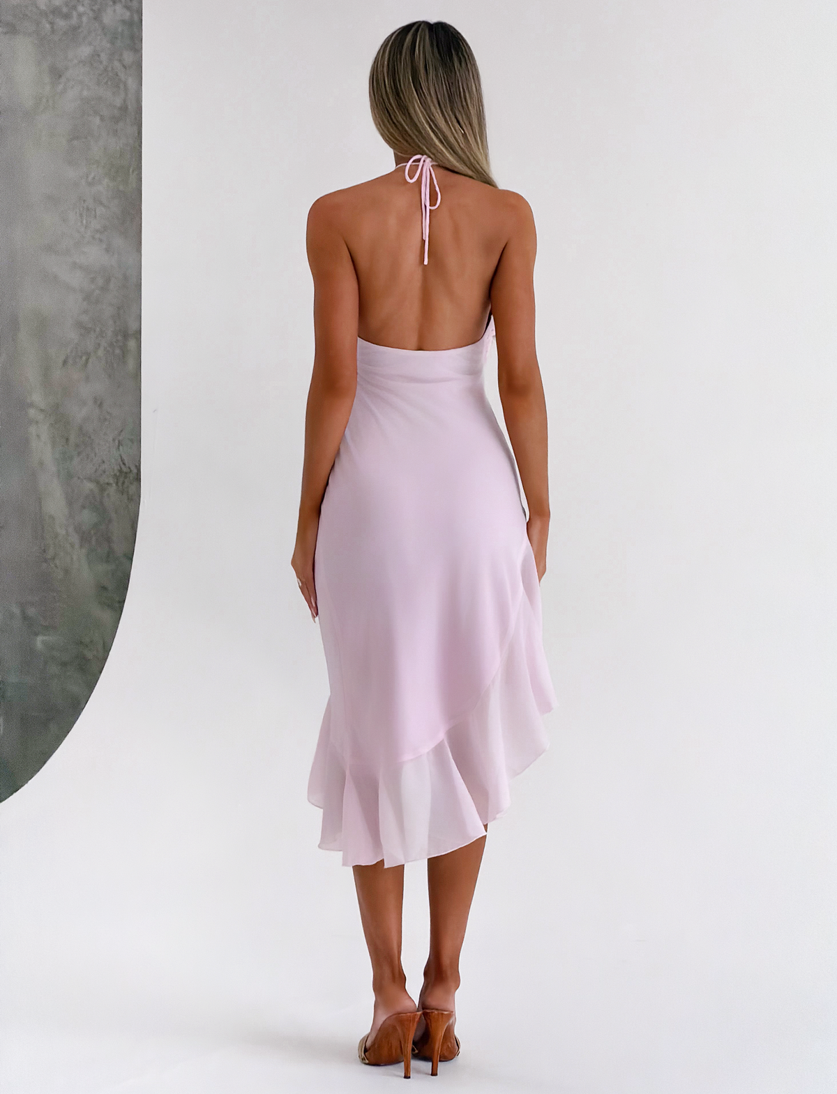 A woman with long blonde hair stands barefoot on a white floor, facing away, wearing the TIGERMIST ELARINA MIDI DRESS - PINK with ruffles, halter-neck, and asymmetrical hem. The background is soft white and grey.