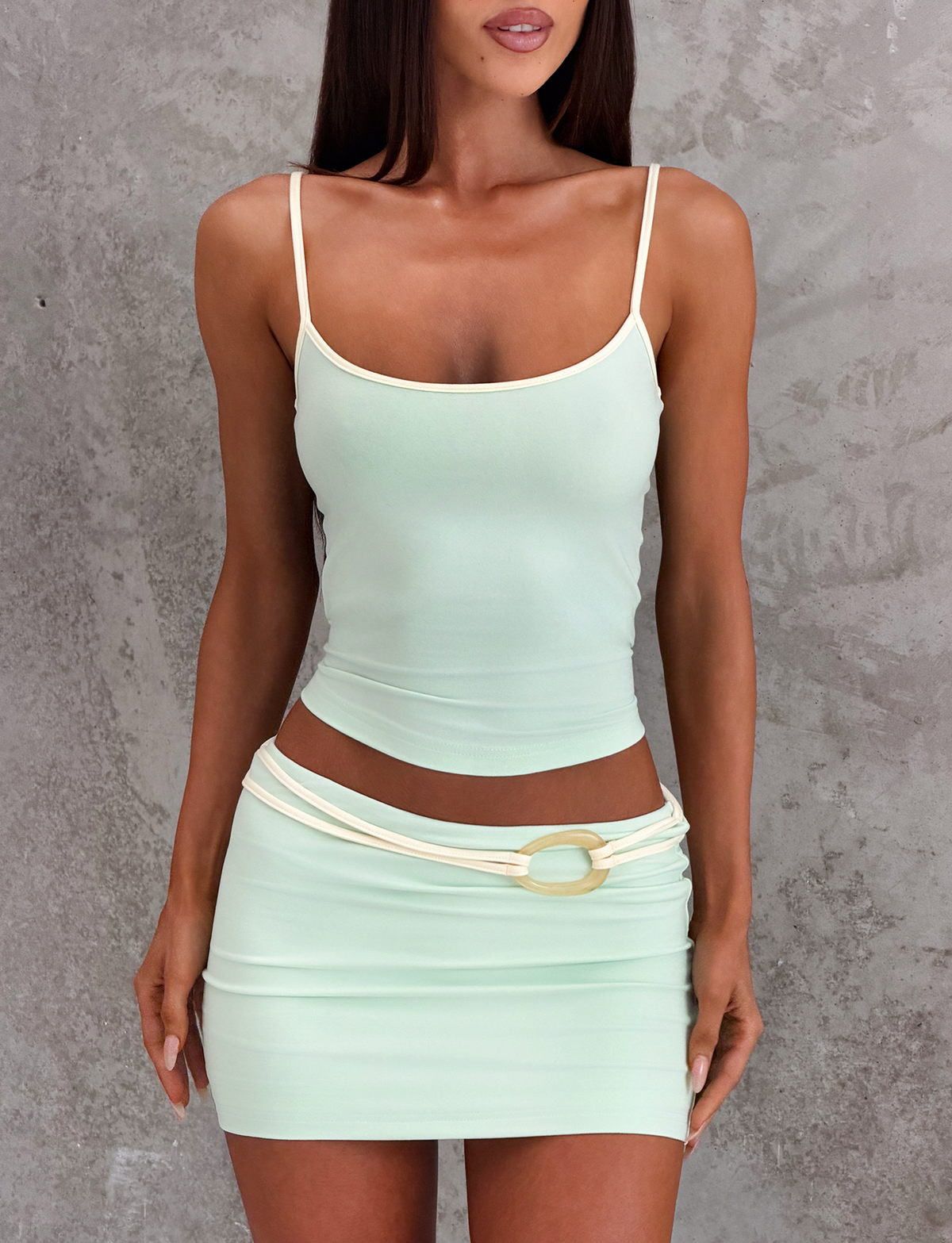 A woman models the TIGERMIST GREER TOP in mint green, featuring a scoop neckline and adjustable straps, paired with a matching mini skirt with a decorative belt and gold ring, standing before a textured gray background.