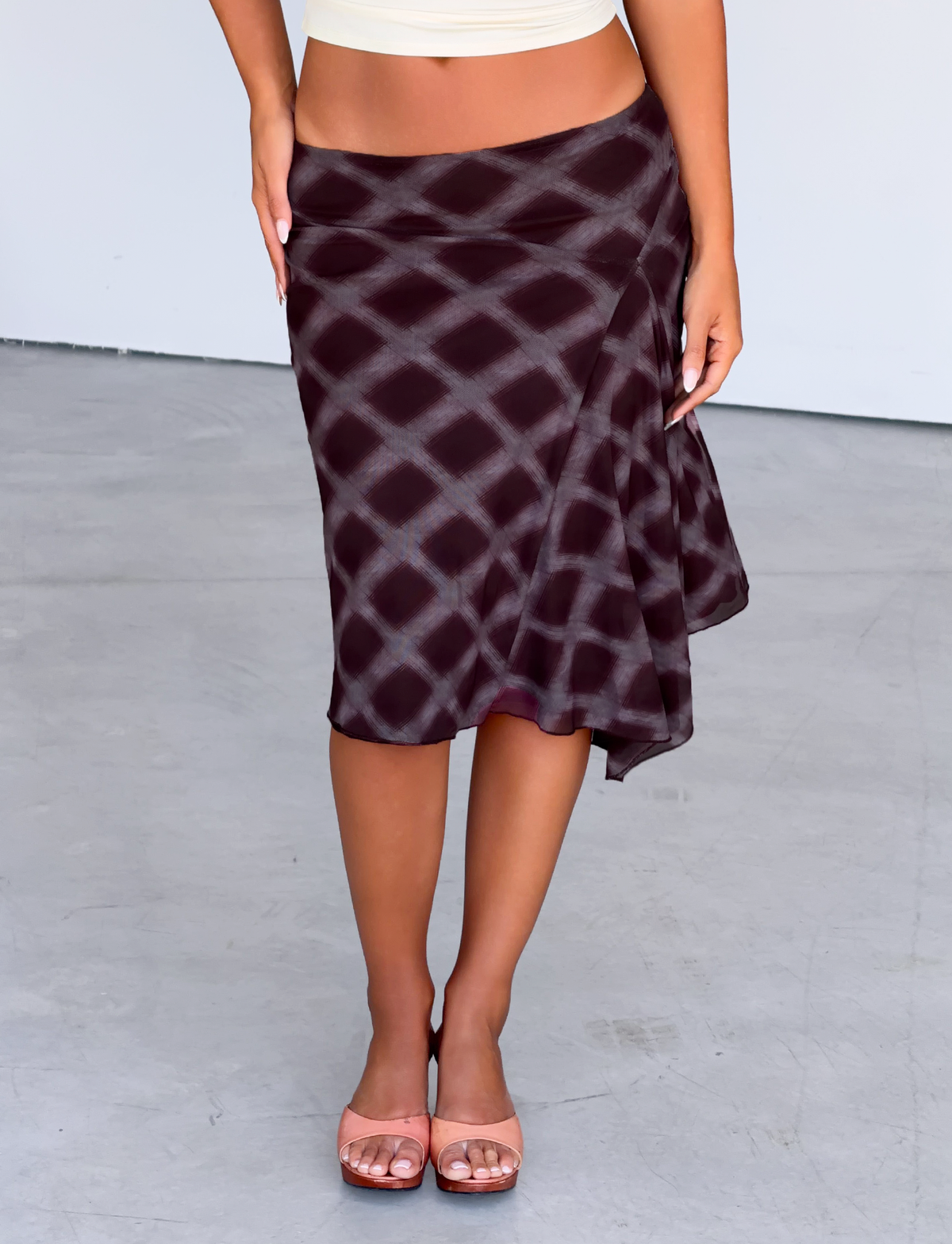 A person wearing the TIGERMIST SAFFRON MIDI SKIRT in Mahogany Check, styled low-waisted with nude heels, stands on a light gray floor, their upper body mostly out of frame.