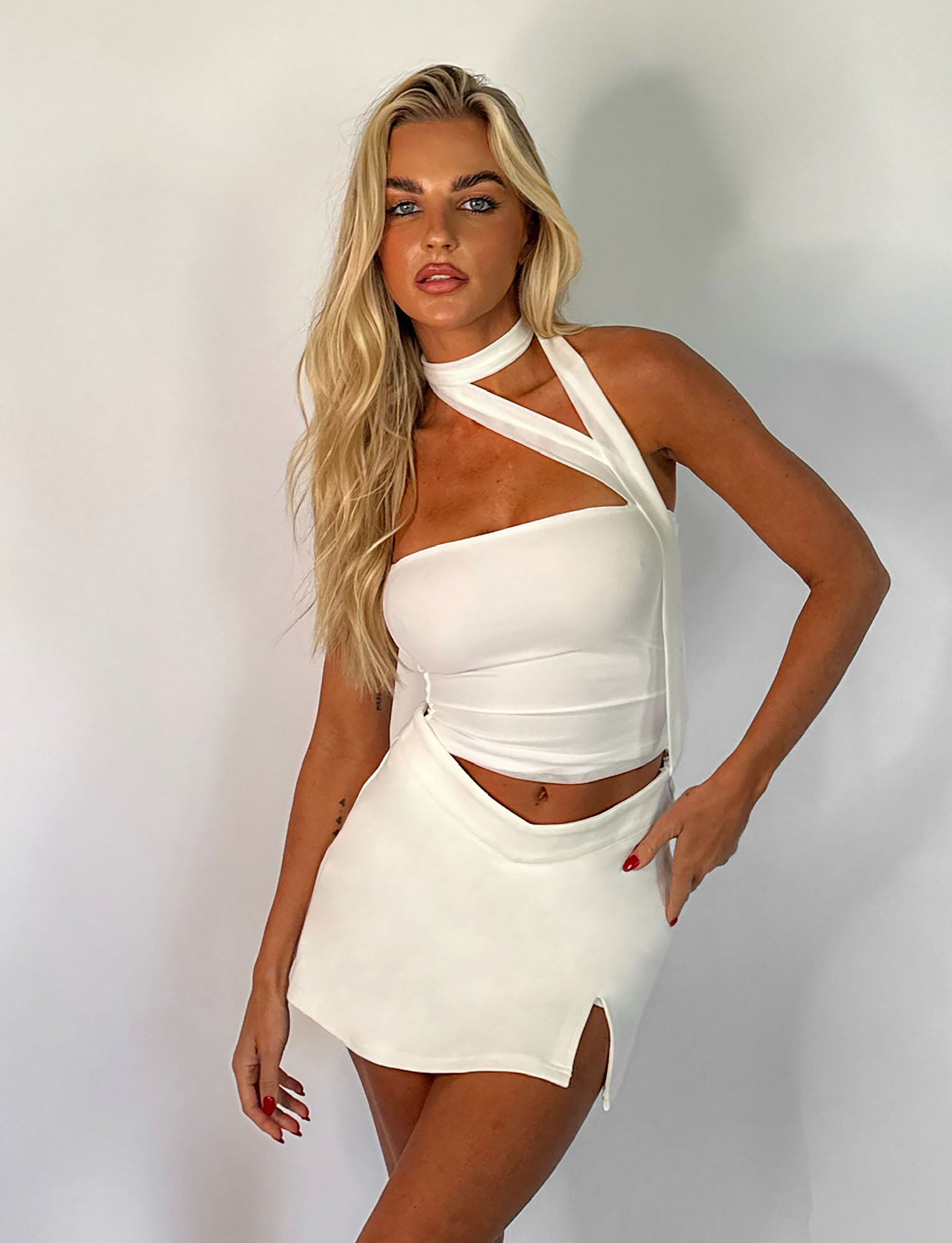 A woman with long blonde hair poses against a white background, wearing a white asymmetrical sleeveless top paired with the DAPHNIE SKORT - WHITE from TIGERMIST. The mini-length skort highlights her red nails as she gazes confidently at the camera.