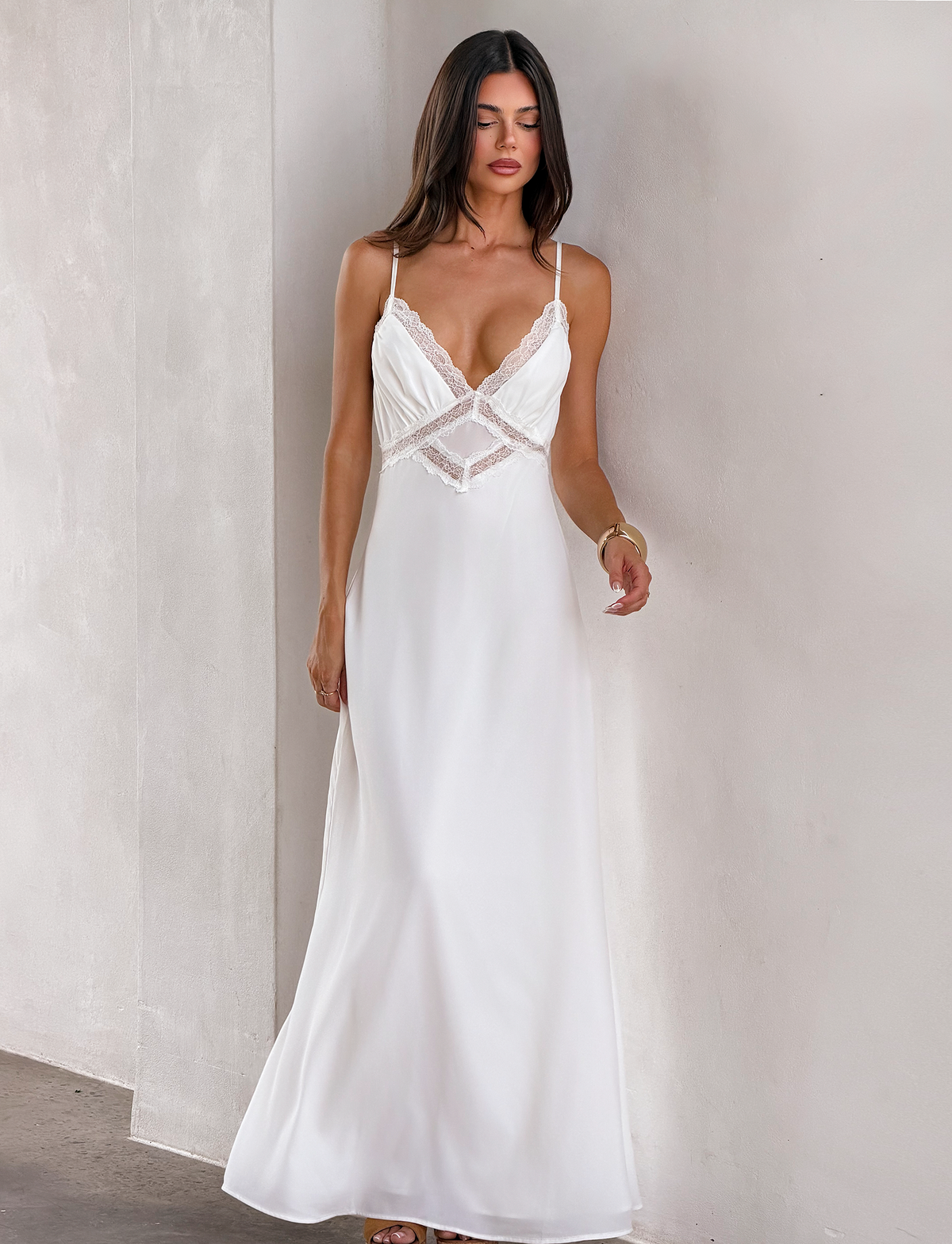 A woman with loose hair and a neutral expression stands against a light wall, wearing the TIGERMIST AISLYNN MAXI DRESS - WHITE—a long slip dress featuring lace trim and thin straps.