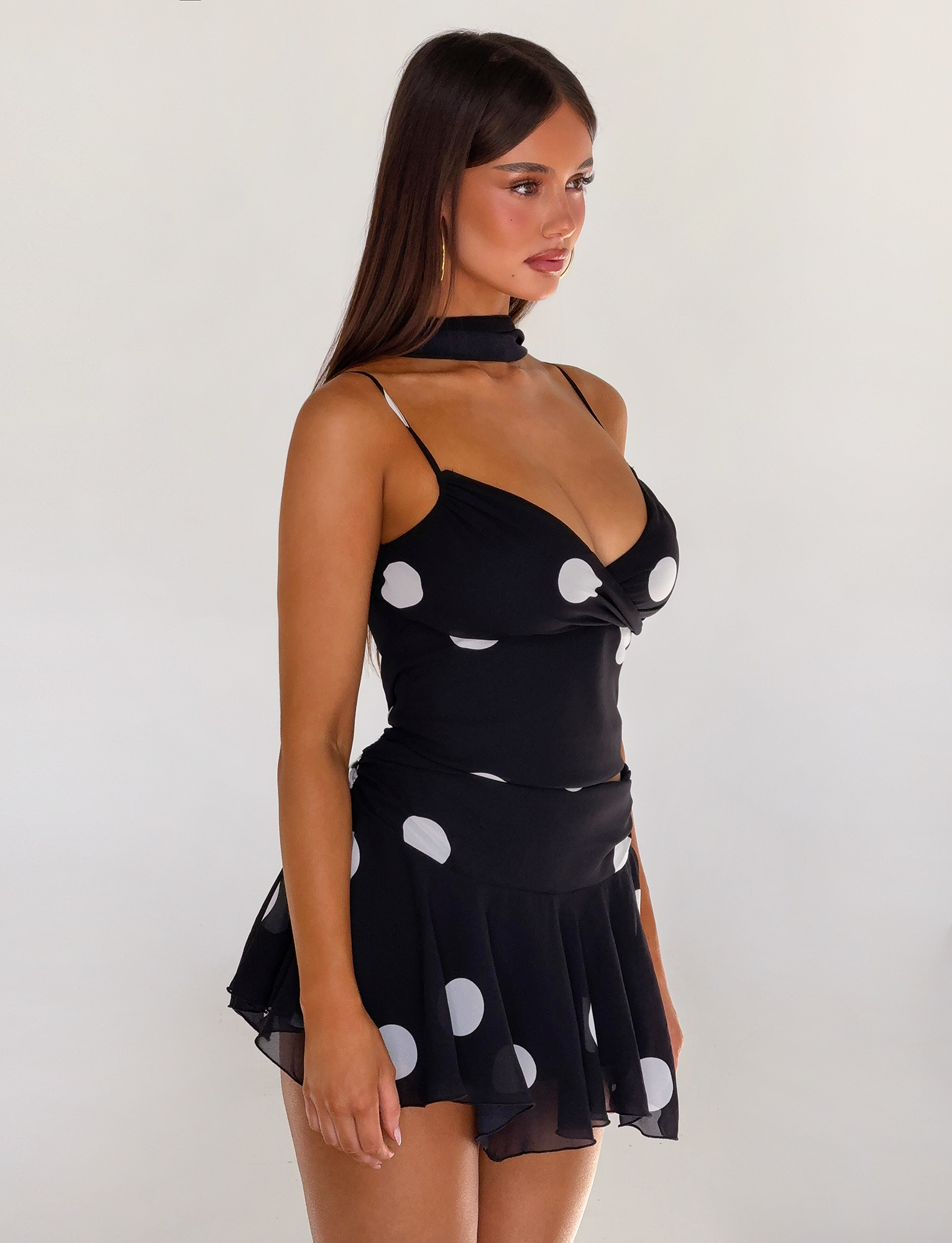 A woman with long brown hair wears the TIGERMIST AYDIN SCARF TOP in black, featuring a polka dot print, deep neckline, adjustable straps, and matching neck scarf, standing against a plain white background.