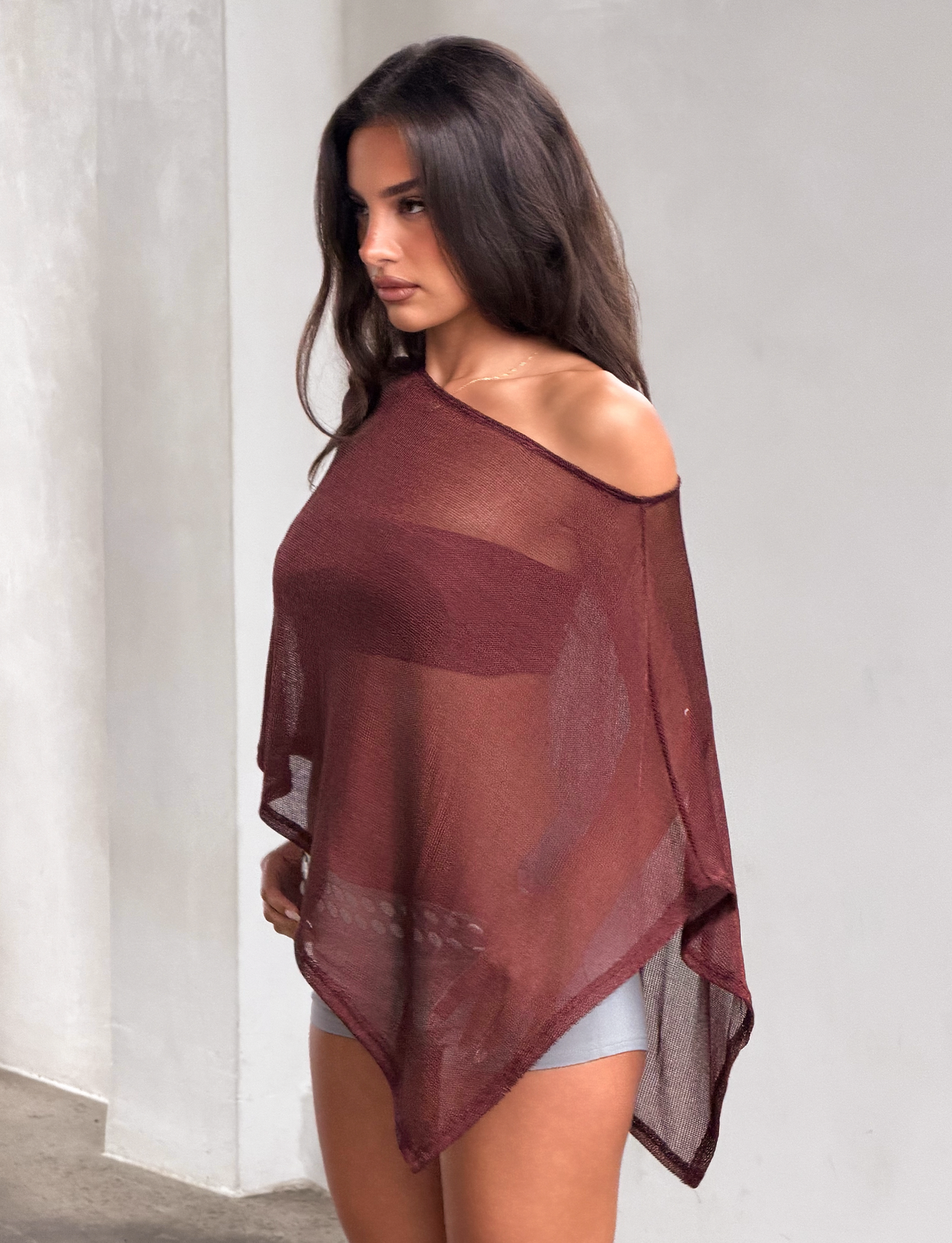 A woman stands indoors with her hair down, looking slightly to the side against a neutral background, wearing the TIGERMIST RAFAELA PONCHO TOP in chocolate over light-colored shorts.