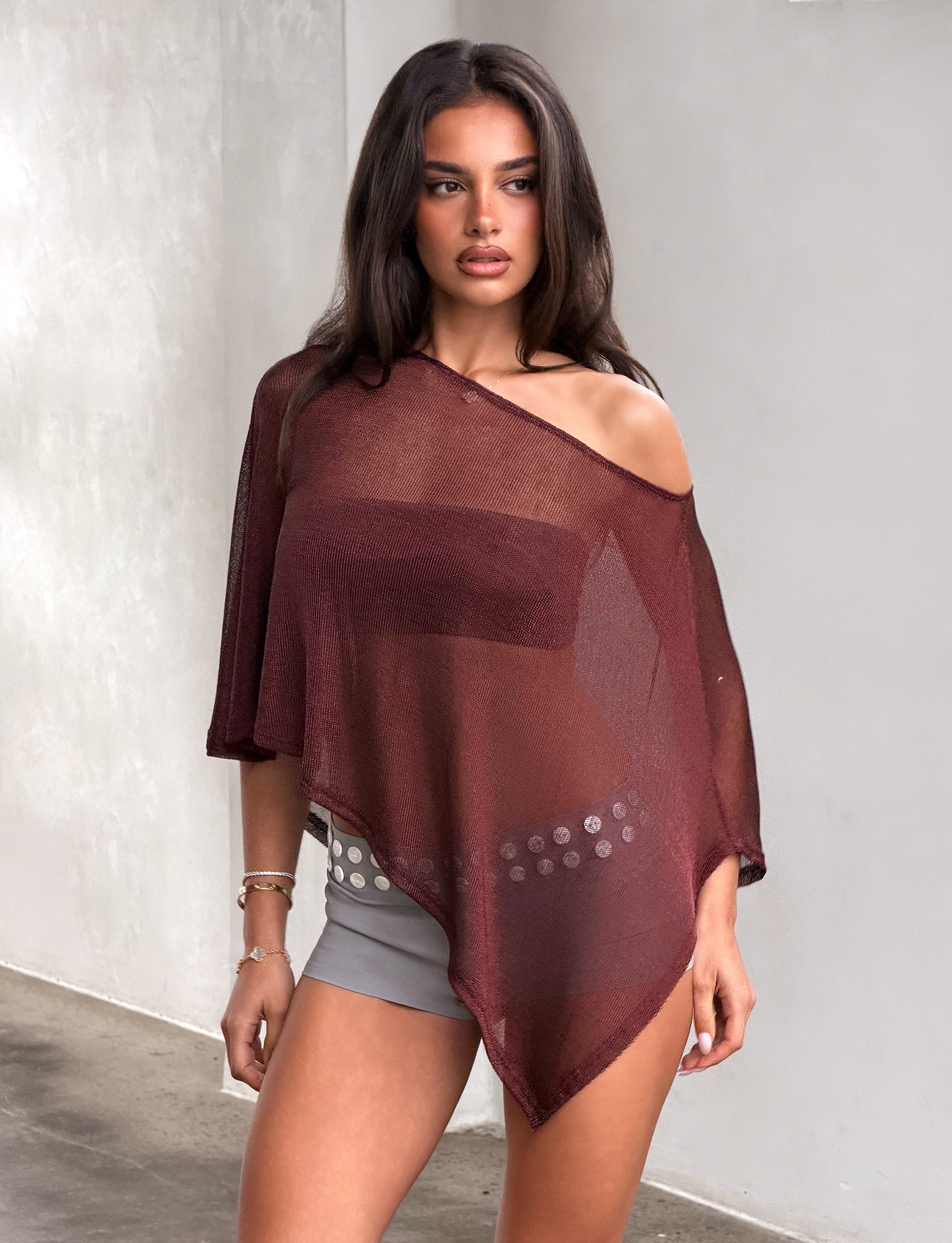 A woman with long brown hair stands against a light-colored wall wearing the TIGERMIST RAFAELA PONCHO TOP in chocolate—a sheer, asymmetrical, off-shoulder style layered over a brown bralette—paired with gray shorts and a decorative belt.