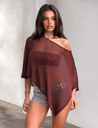 A woman with long brown hair stands against a light-colored wall wearing the TIGERMIST RAFAELA PONCHO TOP in chocolate—a sheer, asymmetrical, off-shoulder style layered over a brown bralette—paired with gray shorts and a decorative belt.