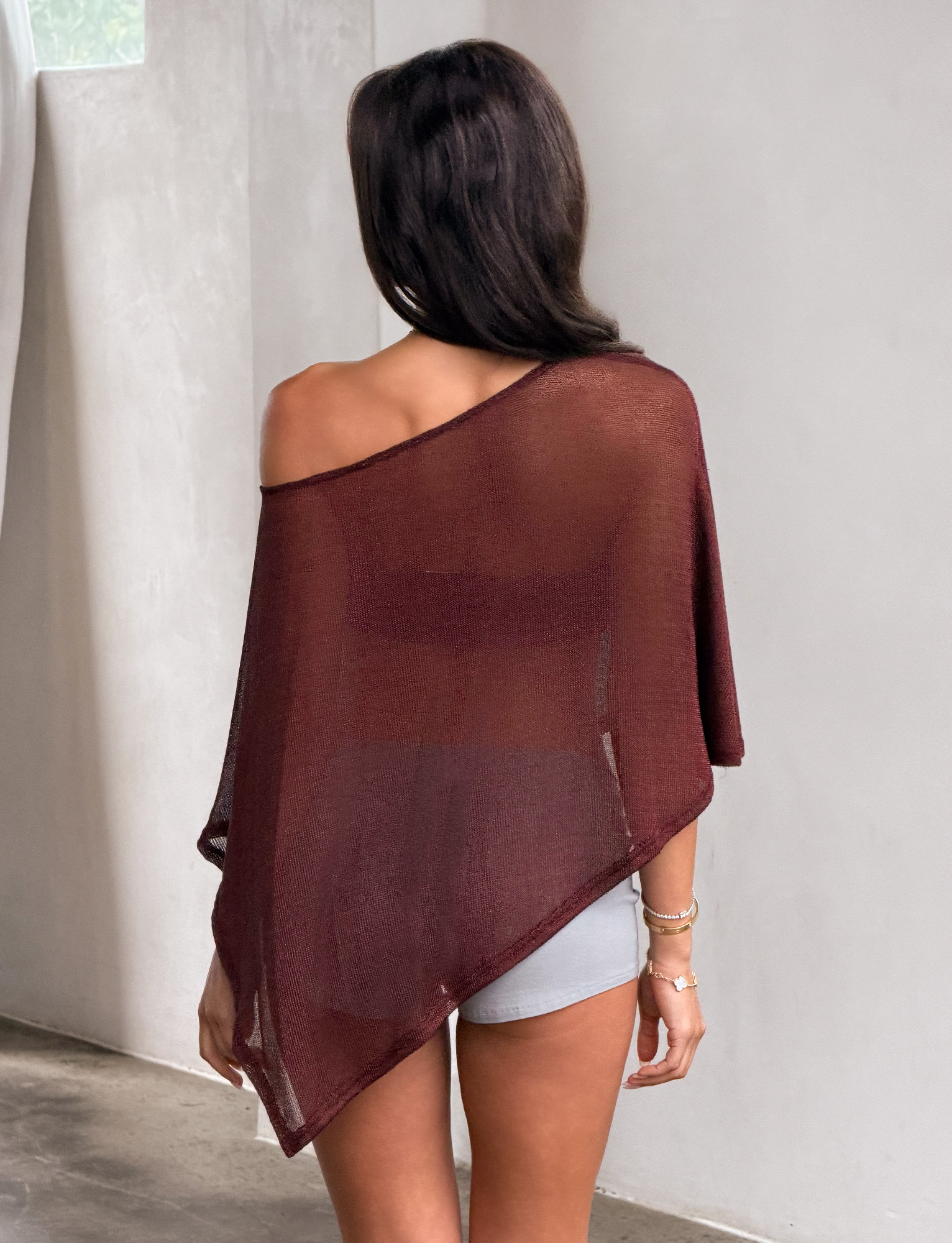 A woman stands indoors with her back to the camera, wearing the TIGERMIST RAFAELA PONCHO TOP in chocolate over white shorts. She has long brown hair and accessorizes with bracelets.