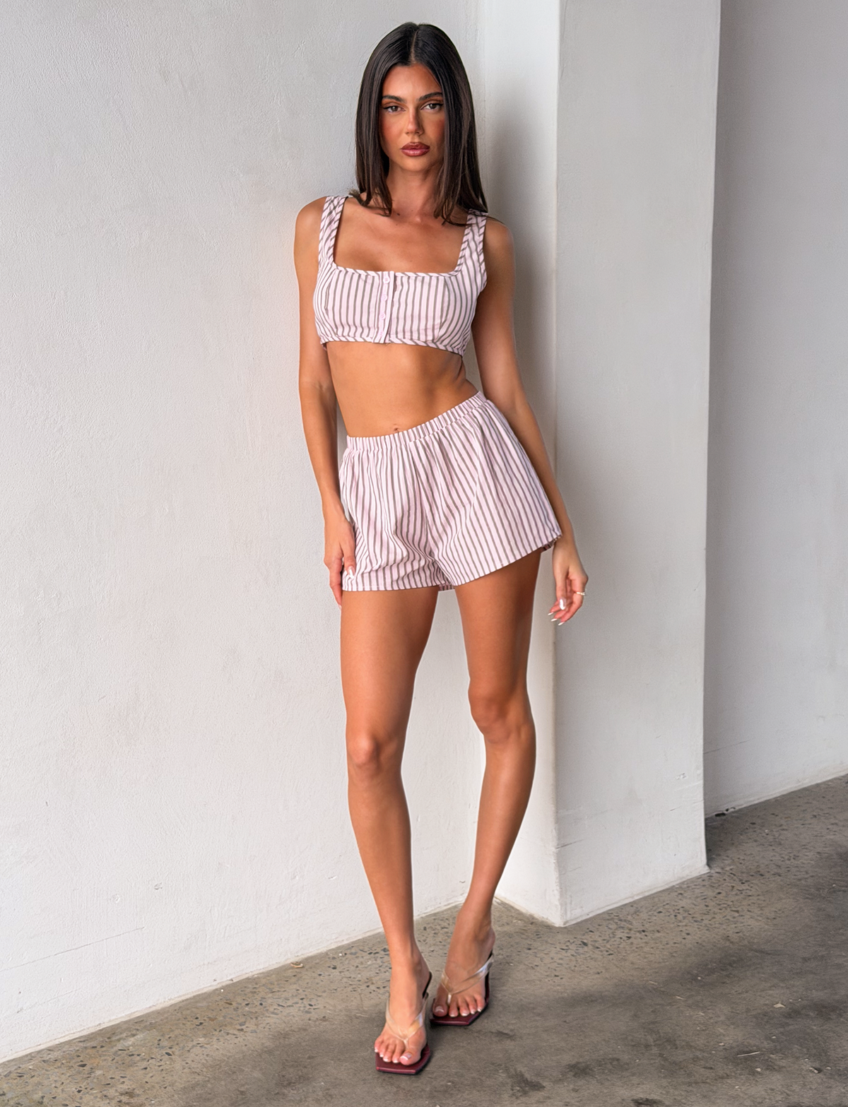 A woman with straight dark hair and a neutral expression stands indoors against a white wall, wearing the TIGERMIST EZRA SHORT - PINK STRIPE set with matching crop top, paired with clear-strap sandals.