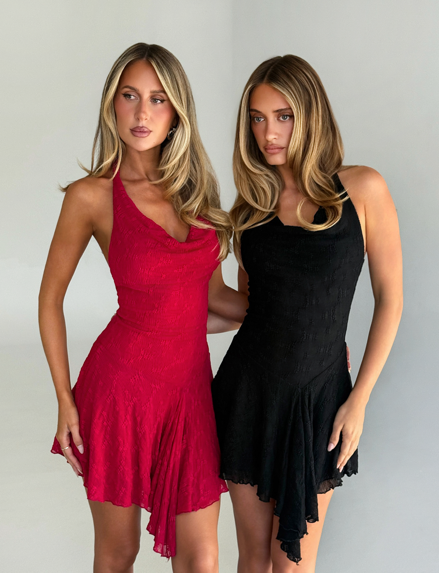 Two women with long blonde hair stand side by side against a light background; one wears the TIGERMIST FIORELLA DRESS - RED LACE, the other a black sleeveless dress. Both have neutral expressions.