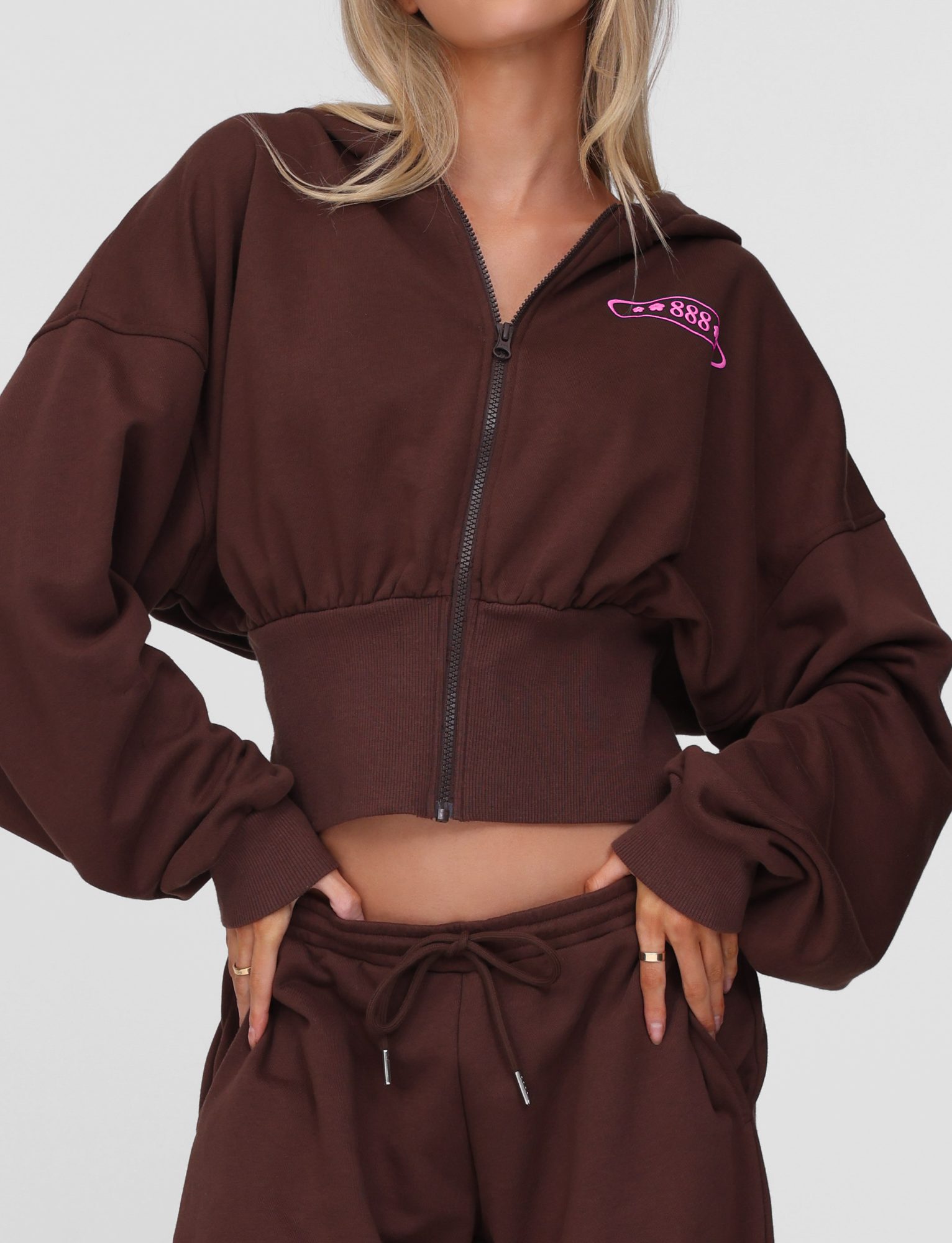 Woman wearing the TIGERMIST MERRYN HOODIE in Chocolate, featuring a pink puff print on the chest, with matching drawstring pants. Her hands are on her hips and her face is mostly out of frame.