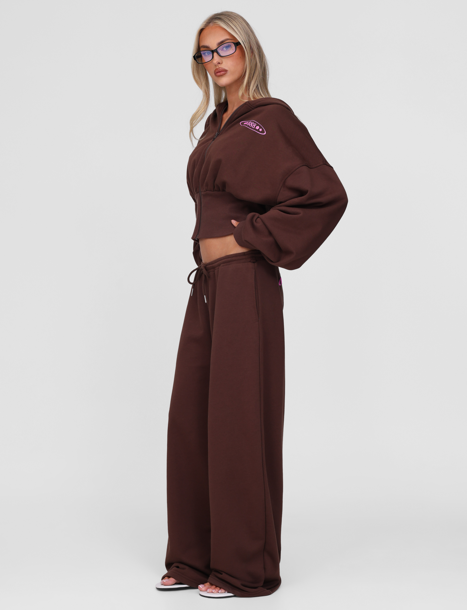 A woman with long blonde hair and glasses stands sideways, barefoot against a white background, wearing the TIGERMIST MERRYN HOODIE - CHOCOLATE and matching wide-leg sweatpants.