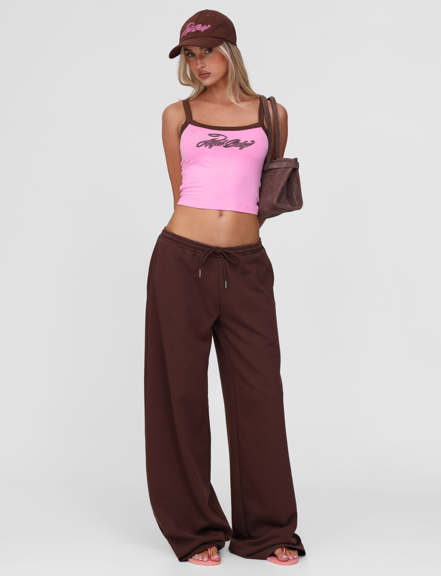 A woman in a pink tank top, TIGERMIST MERRYN TRACK PANT - CHOCOLATE, a matching brown cap, and carrying a brown shoulder bag stands against a plain white background.