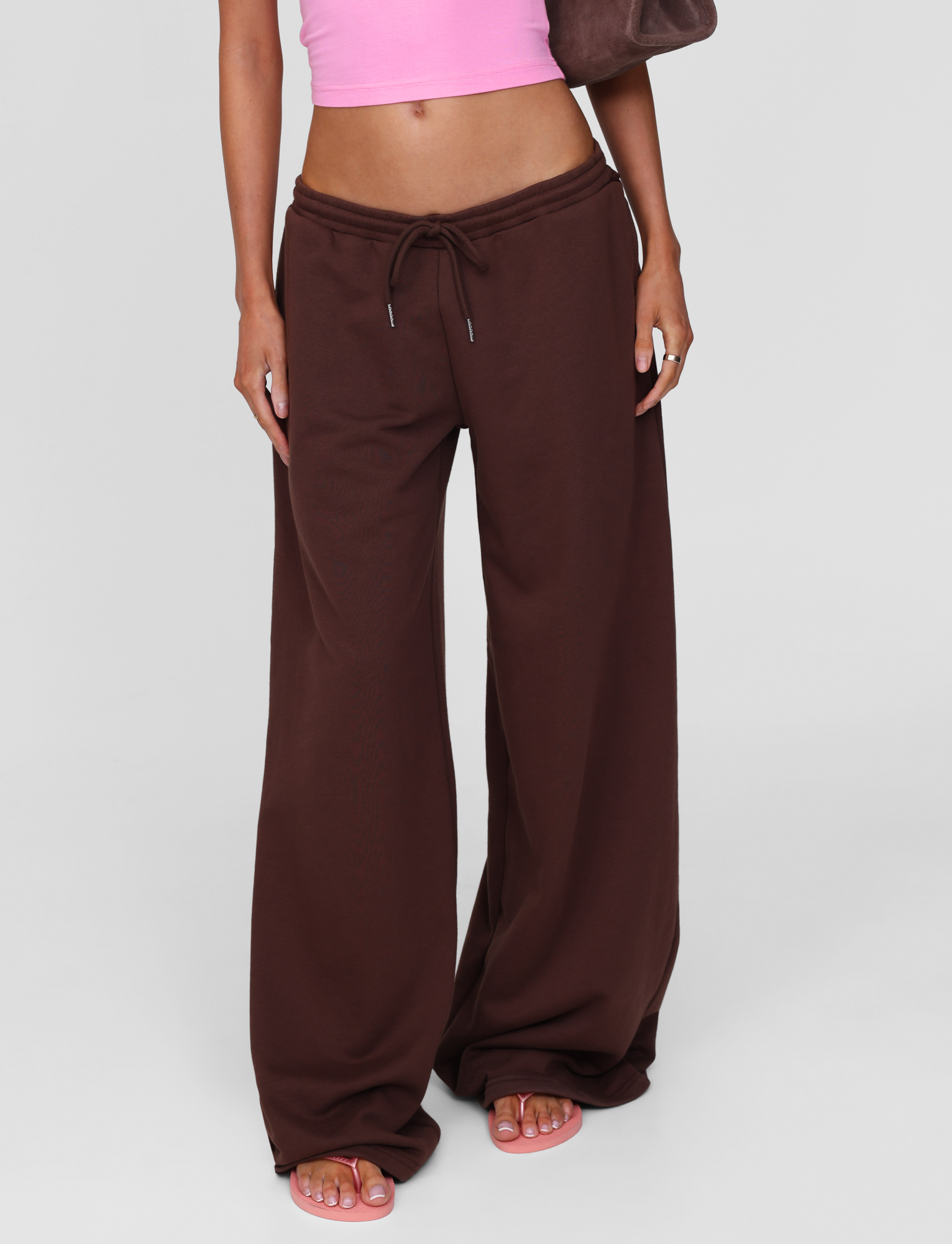 A person wears TIGERMIST's MERRYN TRACK PANT in chocolate with a pink crop top, holding a brown bag. Only their body from shoulders down is visible; barefoot with pink toenail polish.