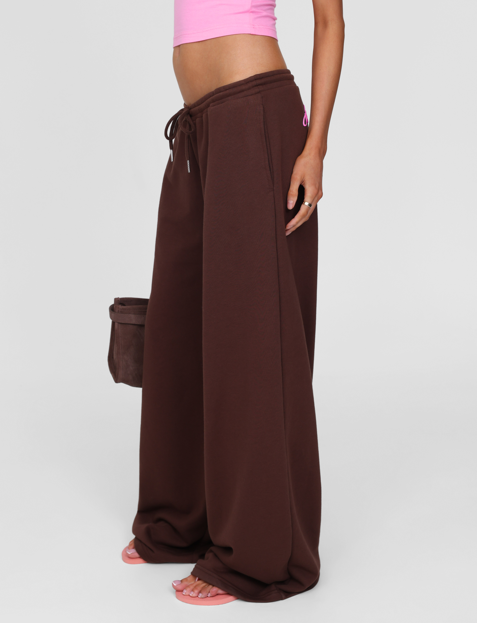 A person is seen from the shoulders down wearing TIGERMIST’s MERRYN TRACK PANT in Chocolate, paired with a pink crop top, pink sandals, and holding a brown bag, set against a plain background.