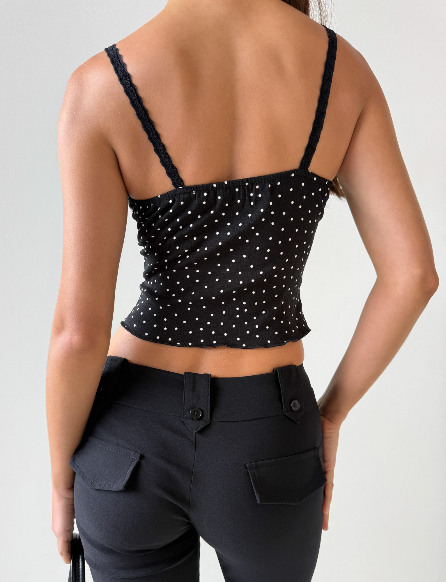 A person is shown from the back wearing the BAILEE CAMI TOP - BLACK POLKA by TOP ONE LADYS FASHION PTY LTD, featuring white polka dots and lace trim, paired with black pants against a plain light background.