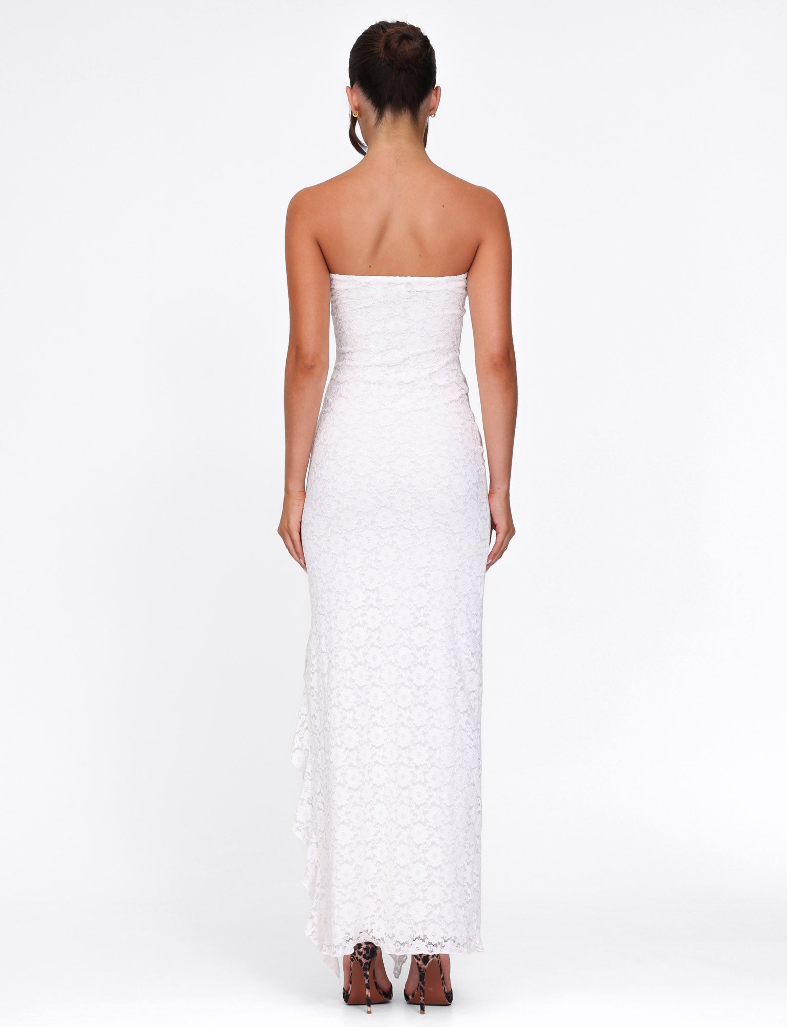 A woman, hair up, is wearing TIGERMIST's THEO LACE MAXI DRESS in white. It's strapless, floor-length with elegant corsetry details and brown heels. She stands against a plain white background.