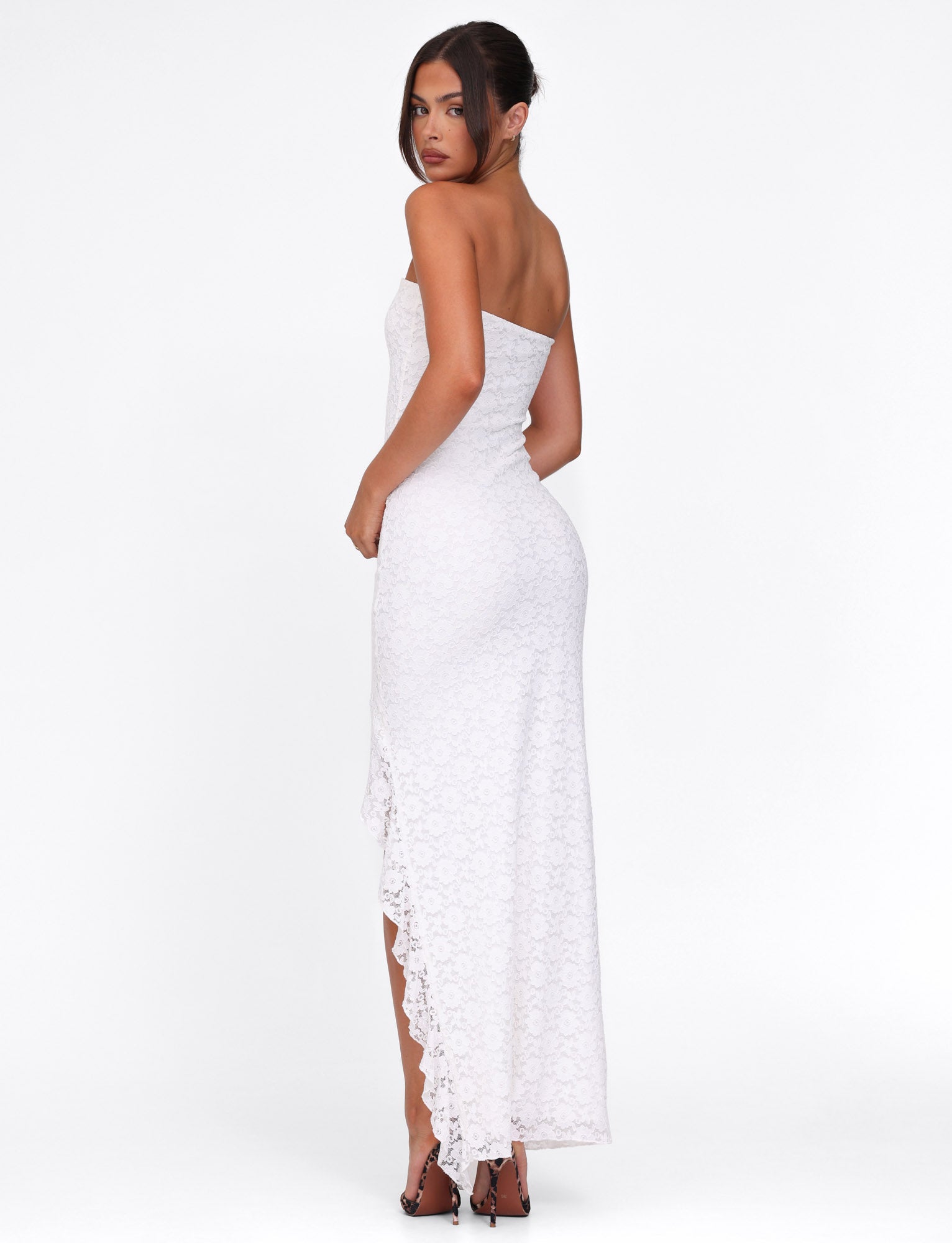 Dressed in the TIGERMIST THEO Lace Maxi Dress - White, featuring a strapless and bodycon design with an asymmetric hemline, a person standing sideways against a white backdrop showcases the gown’s floral lace pattern paired with high heels.