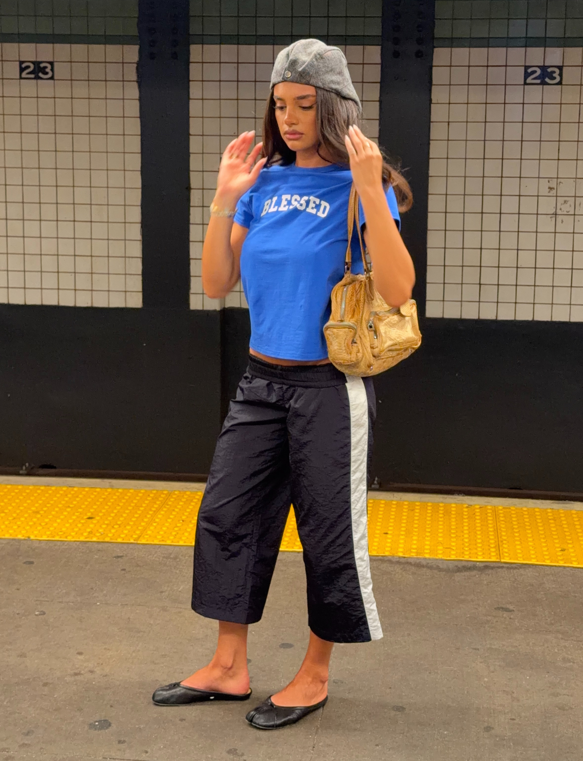 A woman in a blue “BLESSED” shirt, TIGERMIST SOPHIA WINDBREAKER CAPRI – NAVY AND WHITE, black flats, and a gray beret stands on a subway platform, holding a gold handbag.