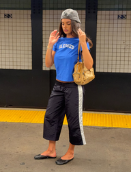 A woman in a blue “BLESSED” shirt, TIGERMIST SOPHIA WINDBREAKER CAPRI – NAVY AND WHITE, black flats, and a gray beret stands on a subway platform, holding a gold handbag.
