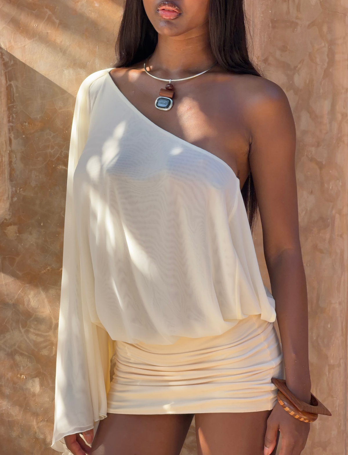 A woman in the TIGERMIST CAMILLE MINI DRESS - CREAM stands against a sunlit wall, accessorized with a necklace and chunky wooden bracelet.