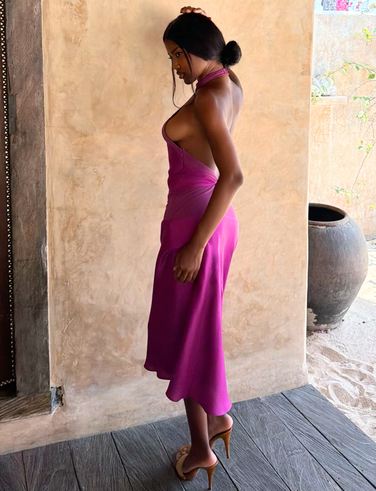 A woman wears the ZAYA MIDI DRESS - MULBERRY by TIGERMIST with nude heels, her hair in a low bun, standing sideways indoors against neutral-toned walls and decor.