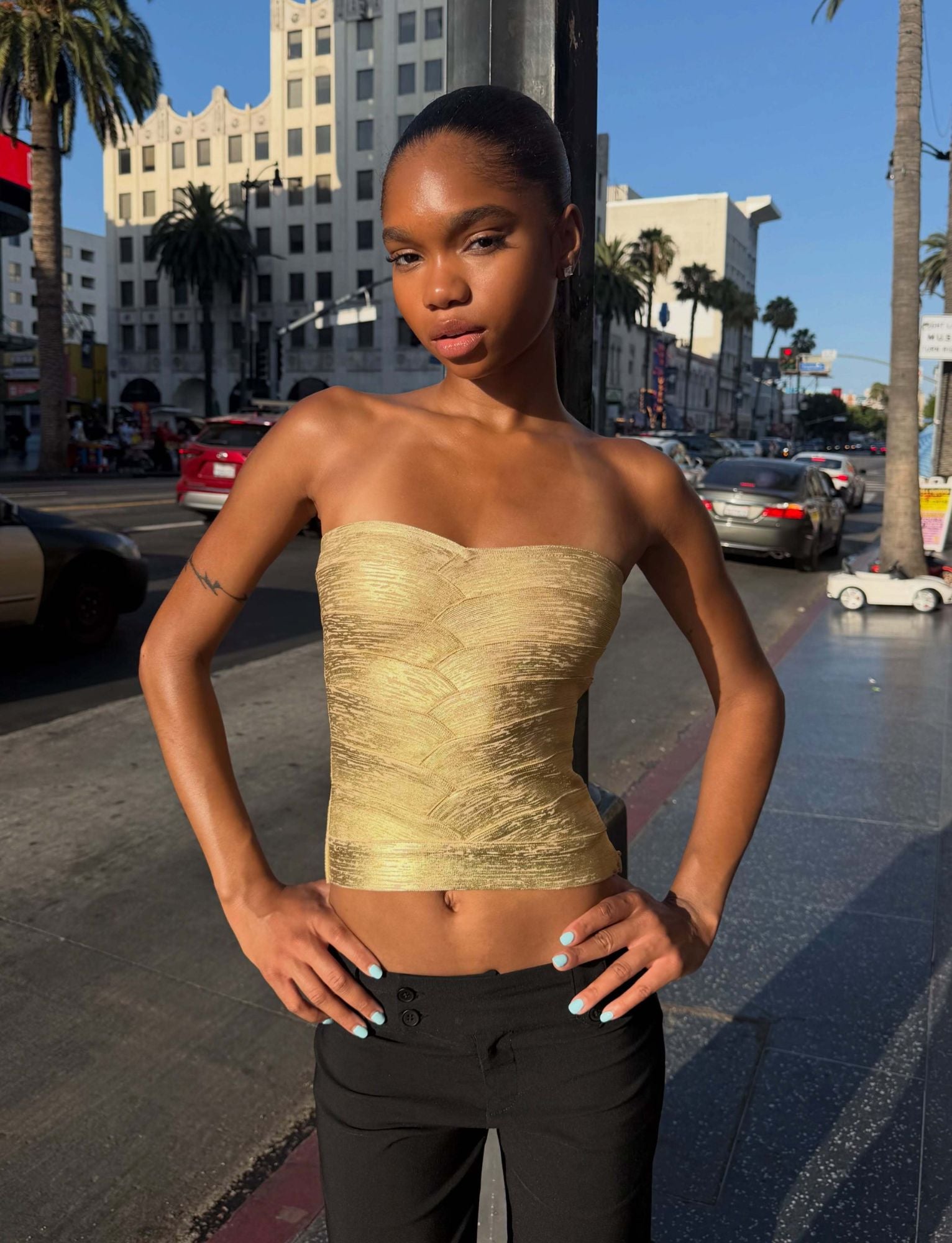 A person poses confidently on a city sidewalk, wearing the TIGERMIST NIYA METALLIC BANDAGE TOP in gold, paired with black pants. Tall buildings, palm trees, cars, and pedestrians can be seen in the sunny urban background.