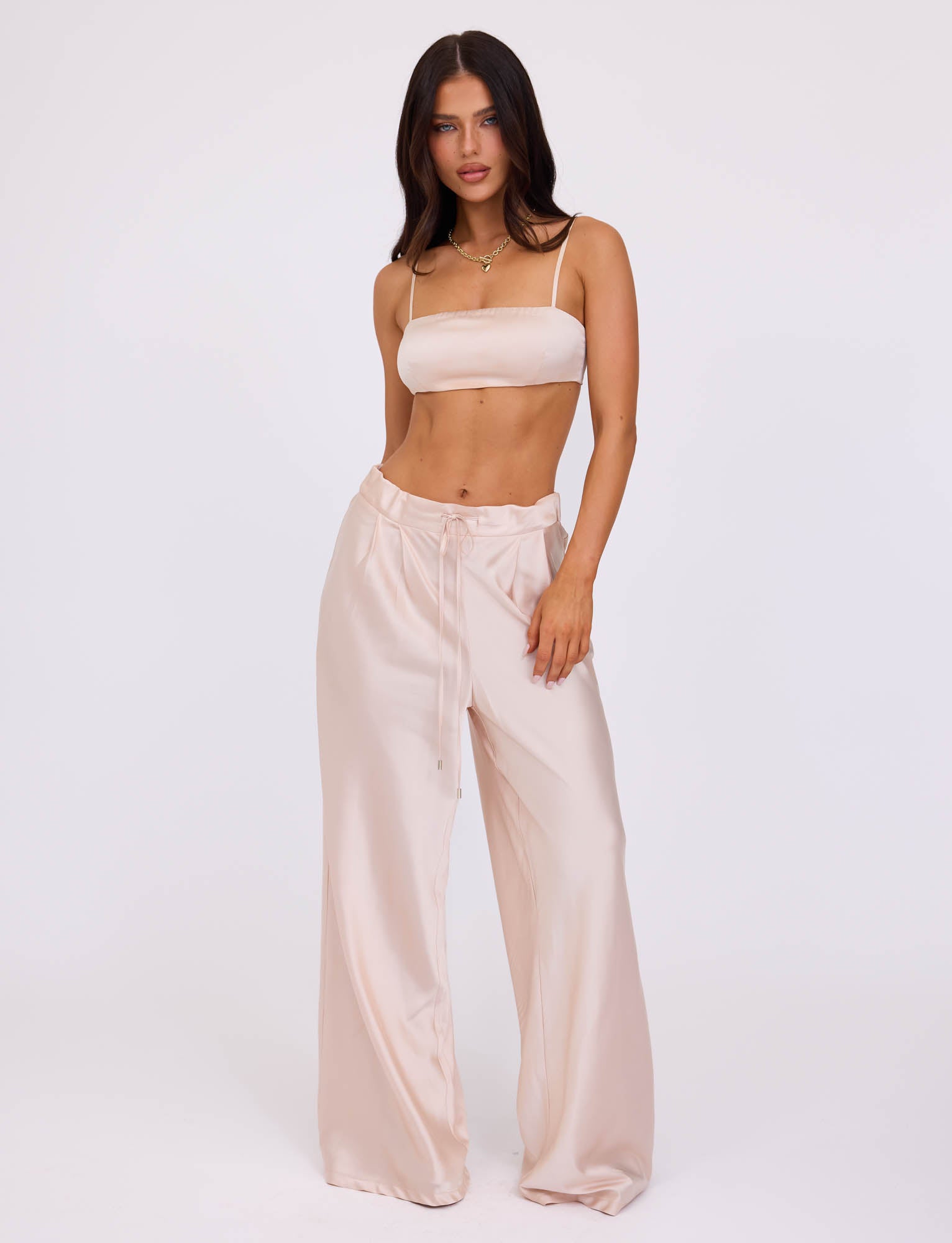 A woman with long dark hair stands against a plain white background, wearing the matching champagne-colored satin crop top and ALINA PANT - WHITE : IVORY from TIGERMIST. She gazes directly at the camera, one hand resting in her pocket, and the other hanging by her side.