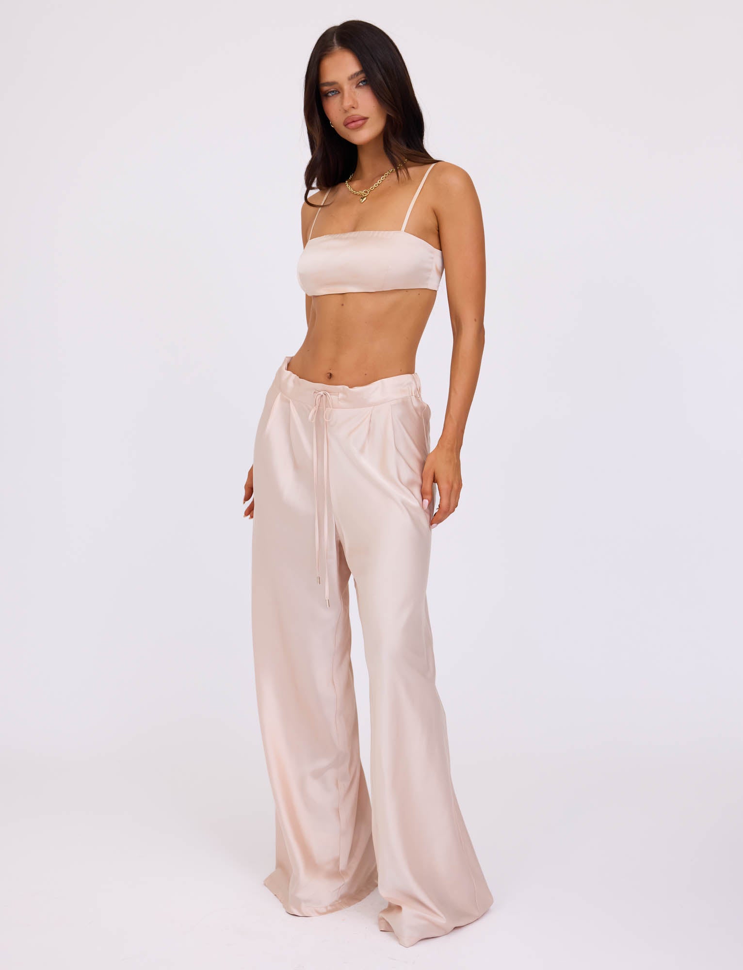 A woman stands against a plain white background, exuding confidence in the ALINA TOP - WHITE : IVORY from TIGERMIST, a satin crop top with thin straps paired with matching high-waisted, wide-leg pants. Her long, dark hair drapes elegantly over her shoulders and is complemented by a delicate gold necklace as she gazes directly at the camera.