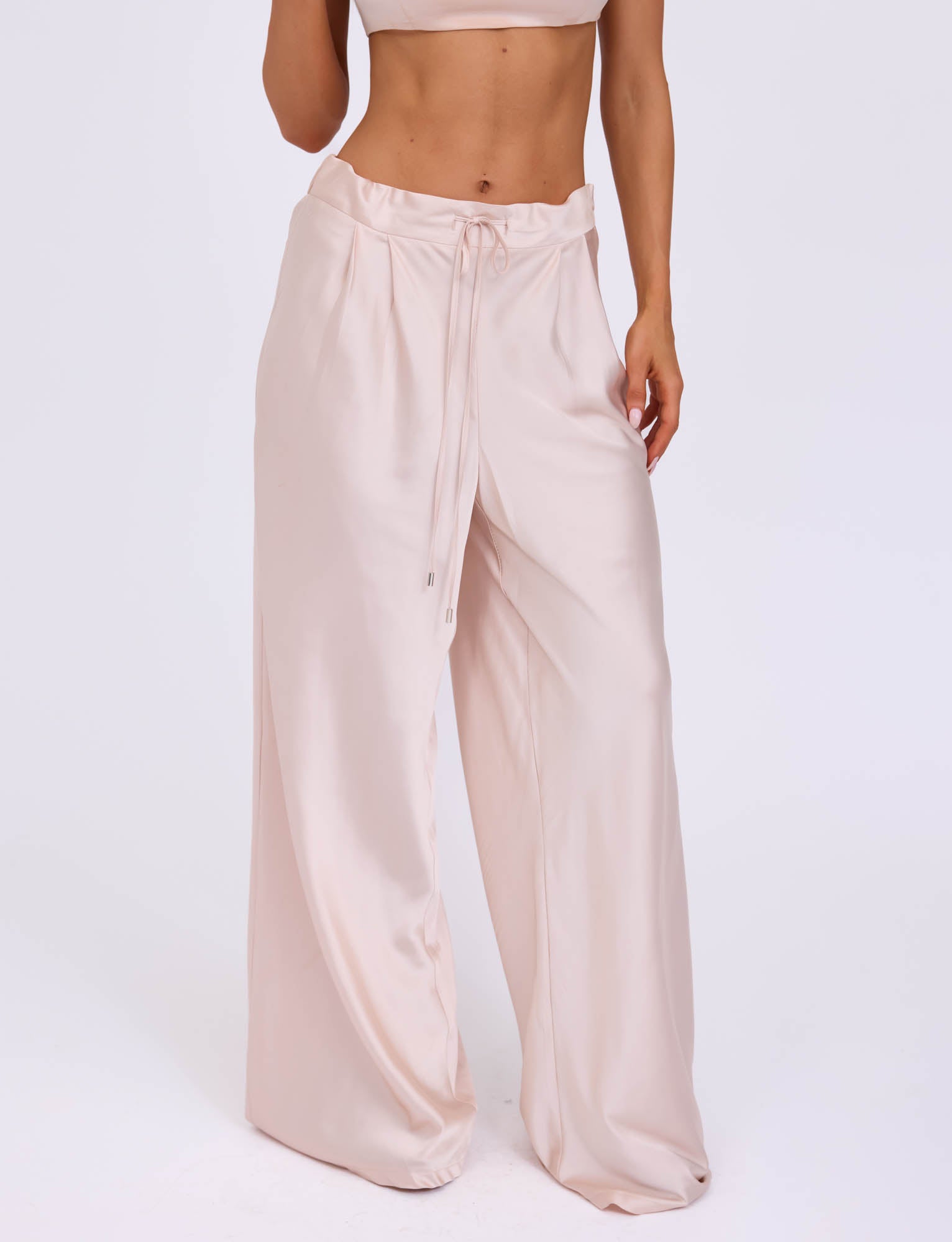 A person is wearing the ALINA PANT - WHITE : IVORY from TIGERMIST. These high-waisted, wide-leg satin pants in light beige feature a drawstring waist. The person pairs them with a cropped top, revealing their midriff, against a plain white background.