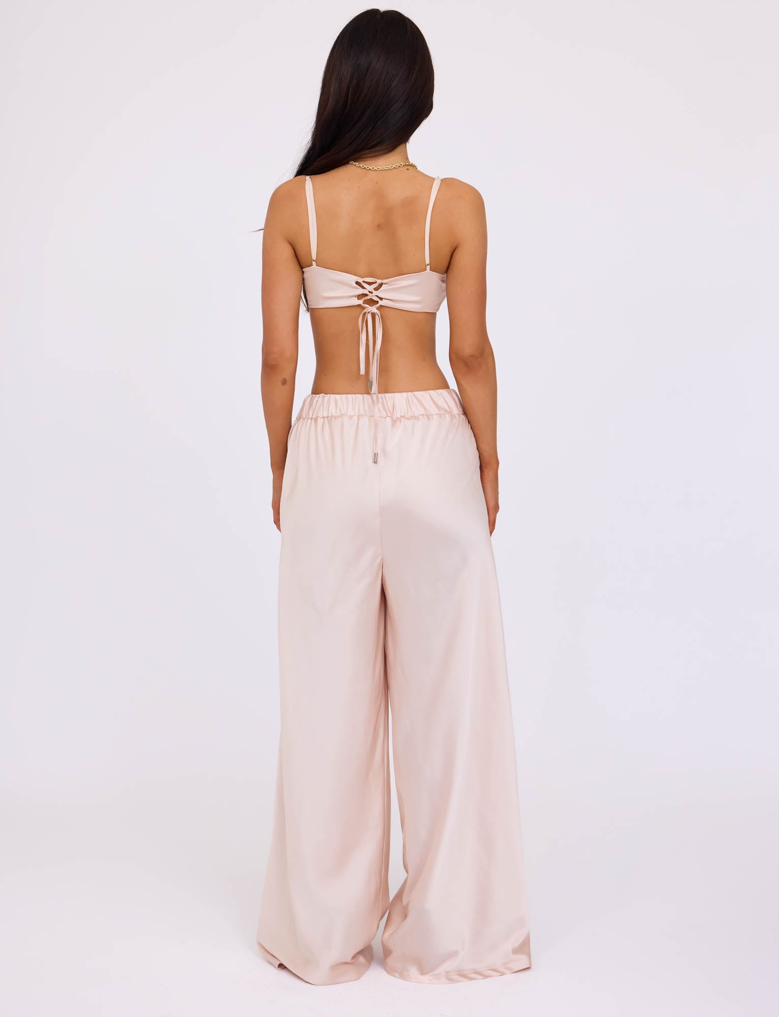 A woman with long, dark hair stands with her back to the camera, highlighting the ALINA TOP - WHITE : IVORY by TIGERMIST, a light pink strappy crop top paired with matching high-waisted, wide-leg pants. The top is designed with criss-cross ties on the back against a plain white background.