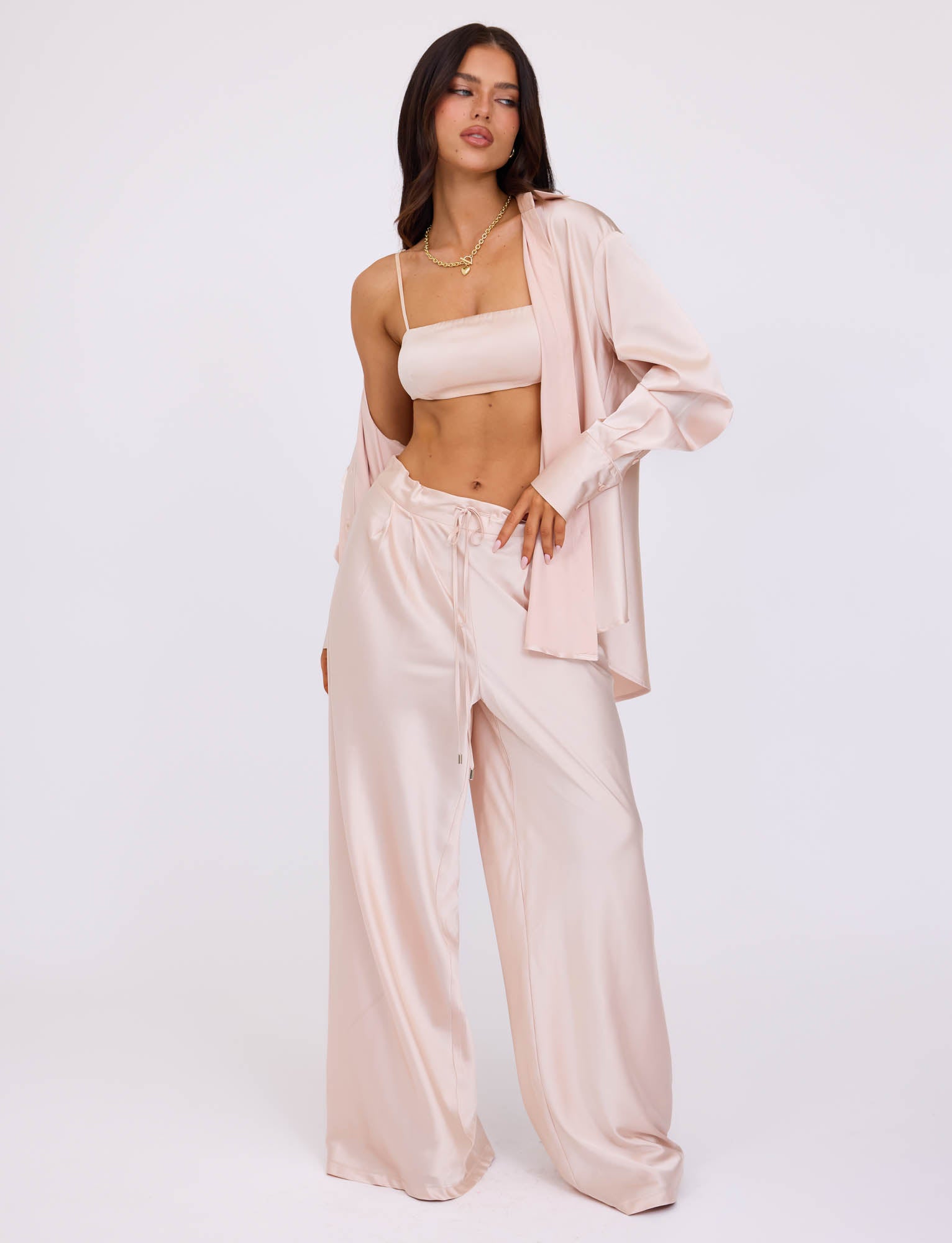 A woman stands against a plain background, wearing a light-colored, silky loungewear set from TIGERMIST. The ensemble includes the ALINA SHIRT - WHITE : IVORY, worn unbuttoned over a cropped camisole and paired with wide-legged pants that feature a drawstring. She has one hand on her hip and the other hanging by her side.