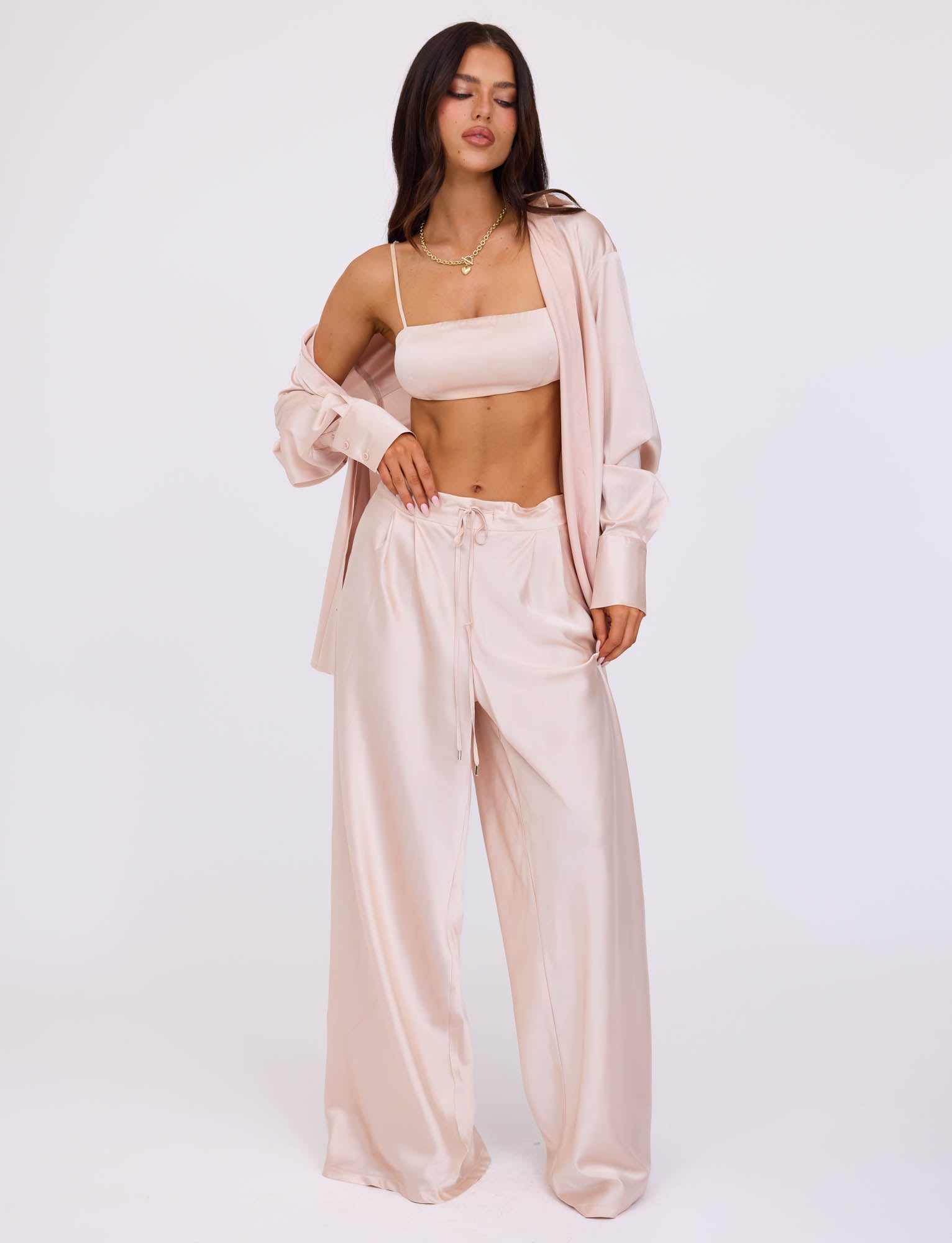 A person stands against a plain white background, dressed in a matching light pink satin ensemble from TIGERMIST. The outfit features a loose, unbuttoned shirt, a cropped top, and the ALINA PANT - WHITE : IVORY with a drawstring waist. The person has long, dark hair and poses with a relaxed expression.