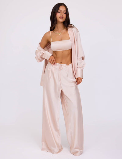 A person with long brown hair stands wearing a light pink silk pajama set from TIGERMIST, featuring a cropped camisole and wide-legged pants, paired with the ALINA SHIRT - WHITE : IVORY draped unbuttoned over their shoulders. They pose with one hand on their hip against a plain white background.