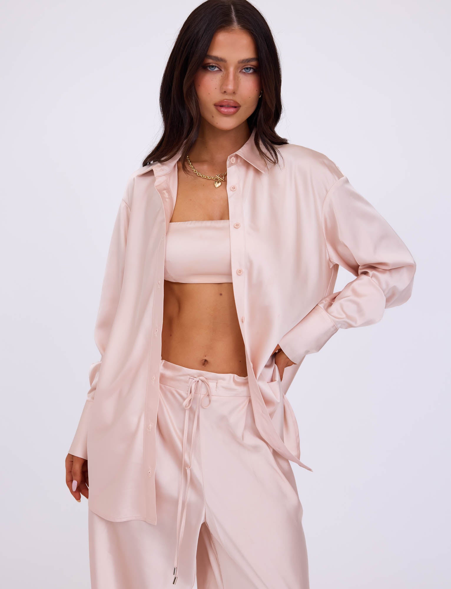 A woman with long, dark hair wears an ALINA SHIRT - IVORY from TIGERMIST, left open over a matching bandeau and drawstring pants. She has a neutral expression, one hand on her hip, and accessorizes with a gold necklace. The background is plain white.